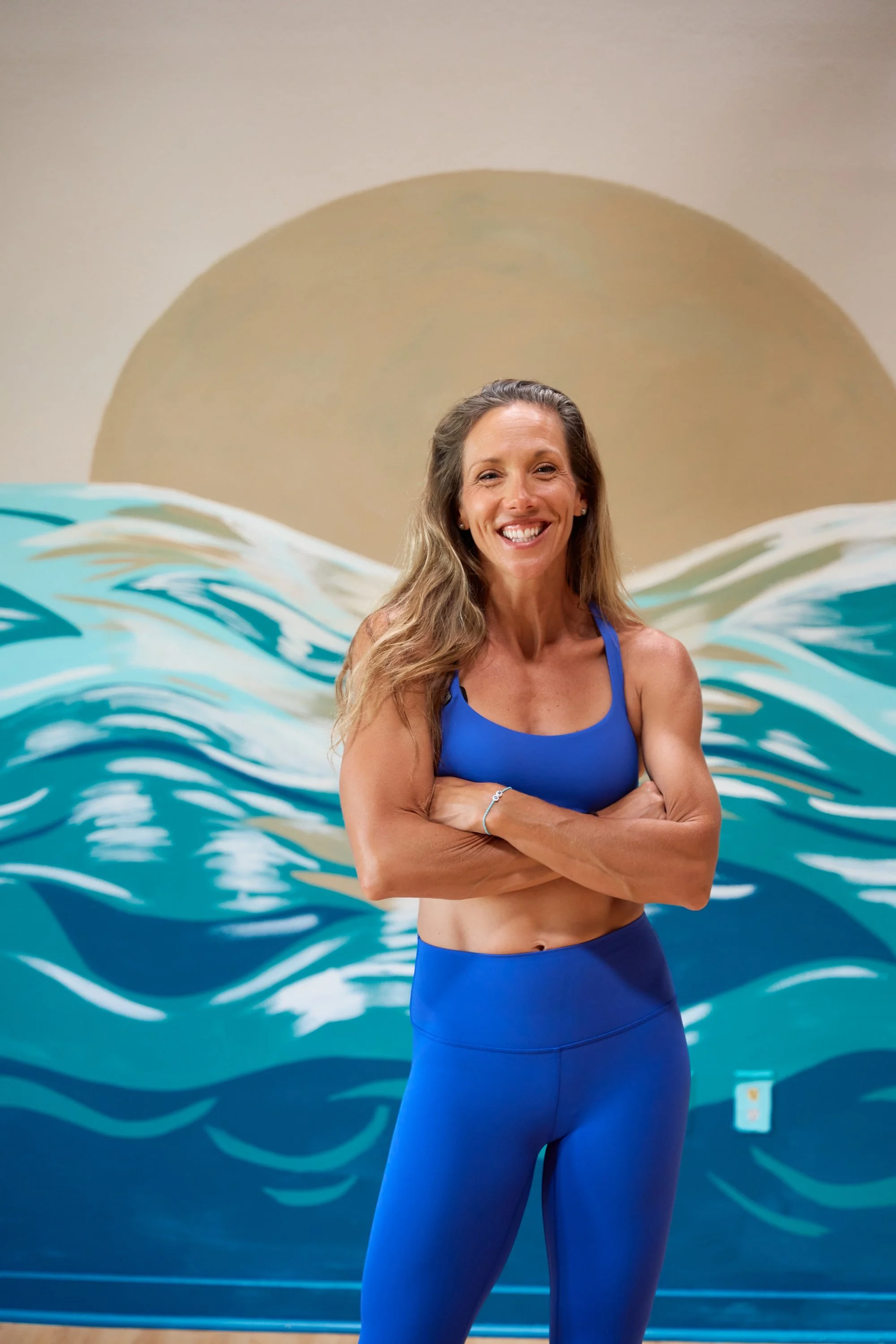 A woman with long wavy hair smiling in front of a mural of waves and a large sun, wearing a blue sports bra and matching leggings.
