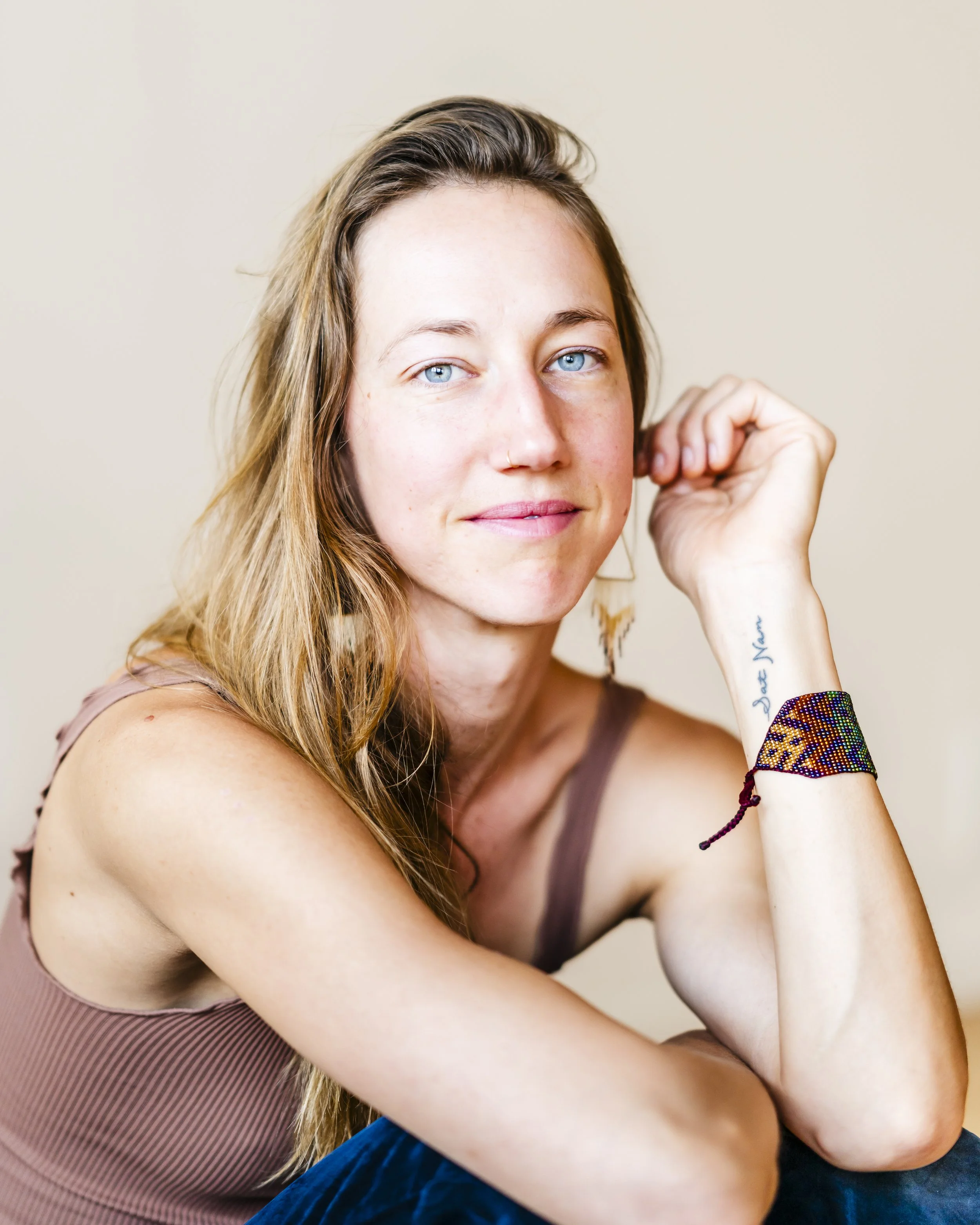 Close-up portrait of a woman with light skin and long wavy blonde hair, sitting against a light beige background, wearing a sleeveless top, with her hand near her face, and wearing colorful beaded bracelets.