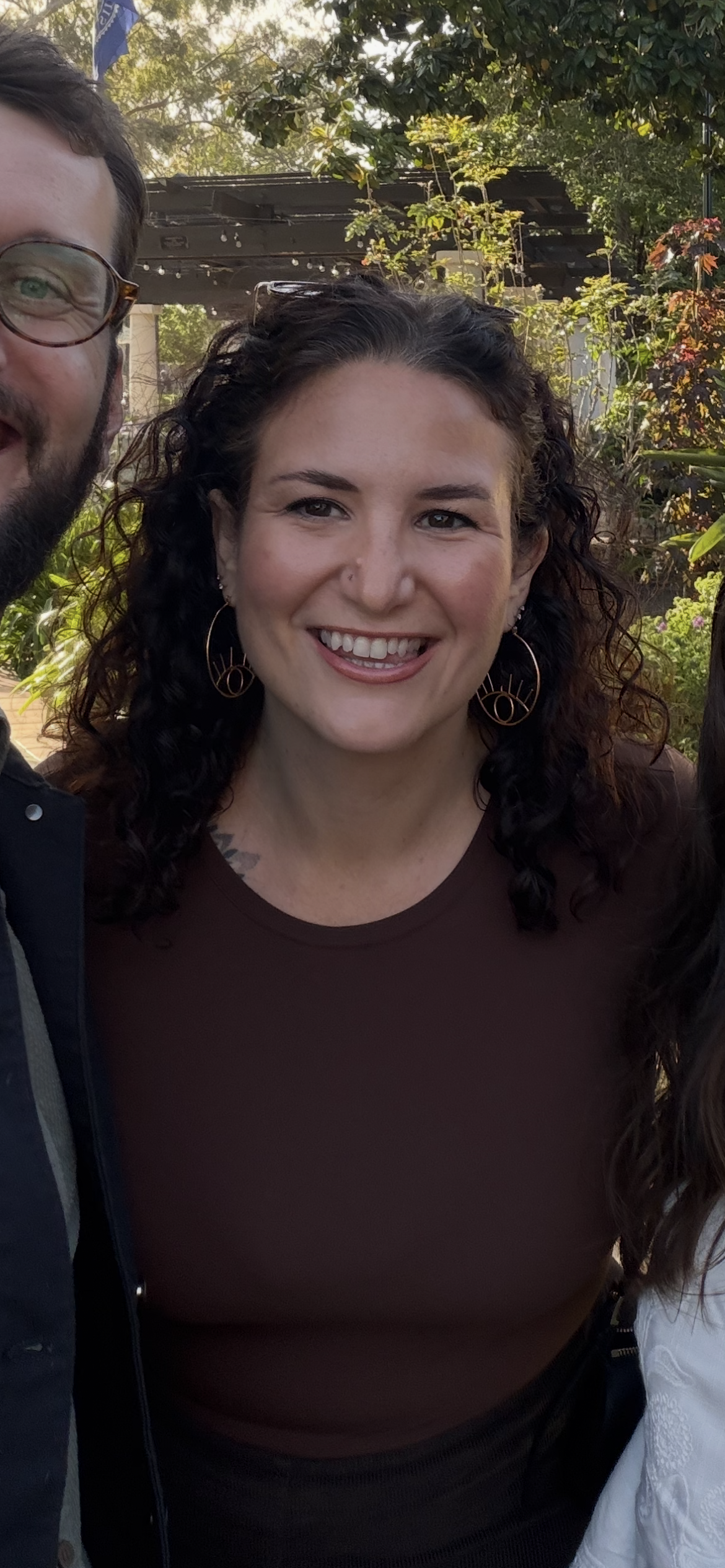 A woman with curly dark hair, hoop earrings, and a maroon top is smiling outdoors during daytime with trees and plants in the background.