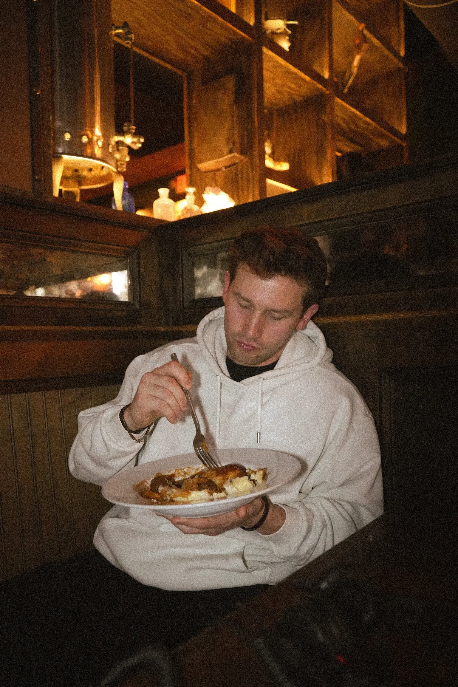 A young man in a white hoodie sitting in a restaurant booth, holding a plate of food and using a fork to eat. The background contains wooden shelves with bottles and utensils.