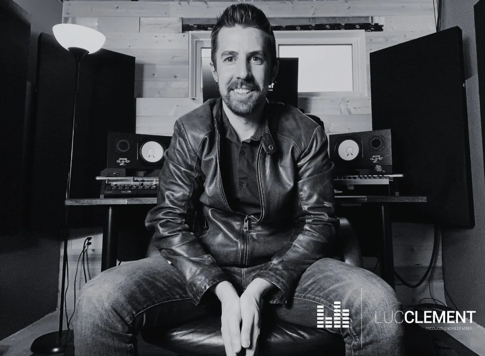 A man with a beard seated in a recording studio wearing a leather jacket, with audio equipment and soundproofing in the background.
