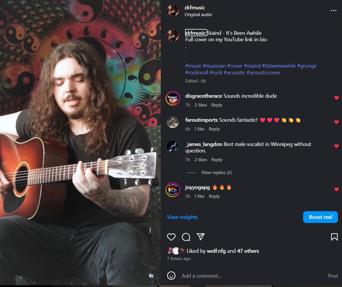 A man with long curly hair and tattoos on his arm is playing an acoustic guitar in front of a colorful, psychedelic tapestry featuring yin-yang symbols.