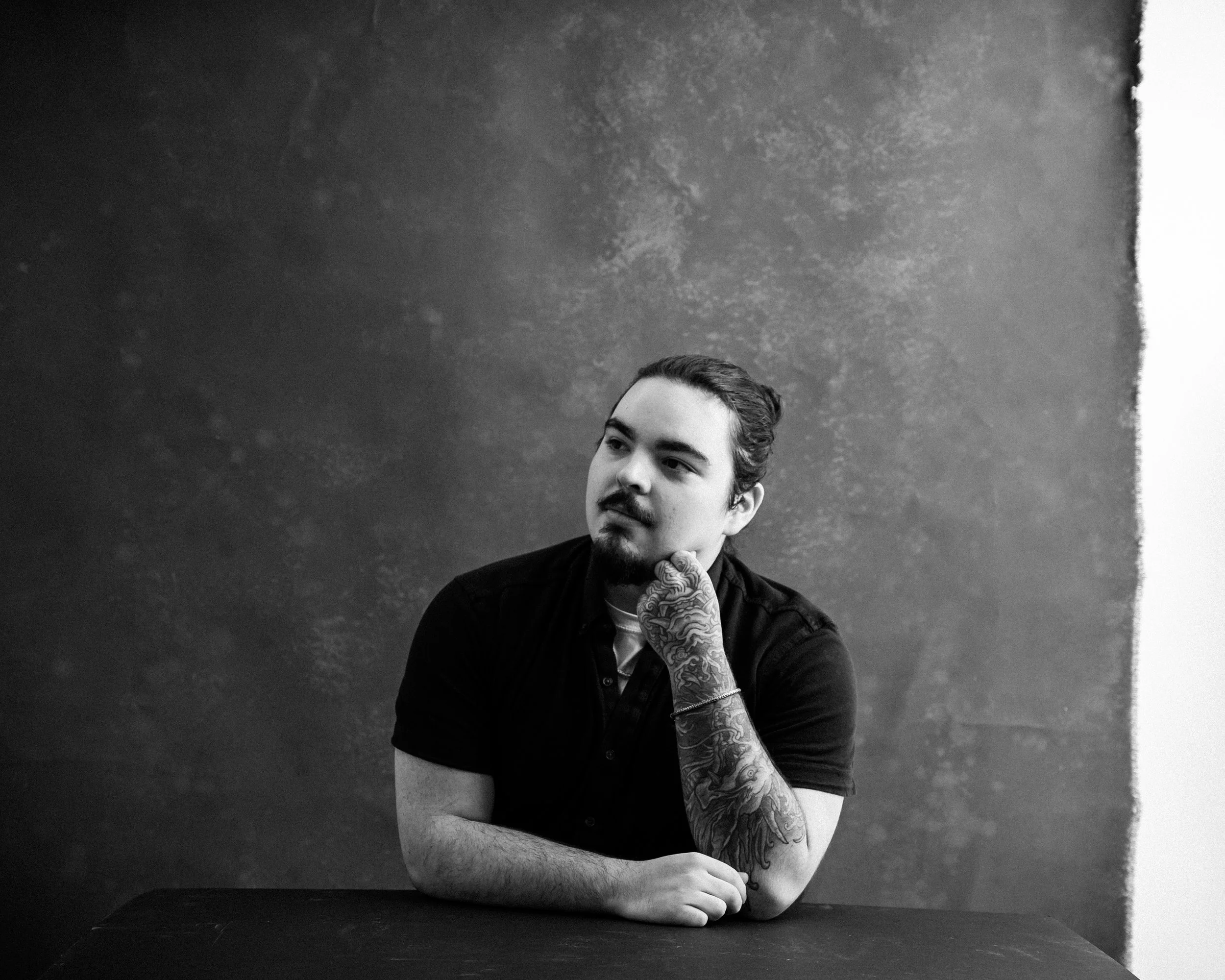 Black and white photo of a man with tattoos on his left arm, resting his chin on his hand, sitting at a table against a textured wall.