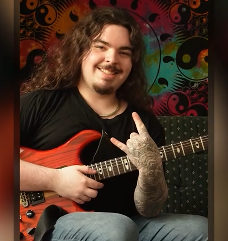 A man with long curly hair, smiling, and showing a rock hand gesture while holding an orange electric guitar. He has a tattooed left arm and is sitting against a colorful, psychedelic tapestry background.