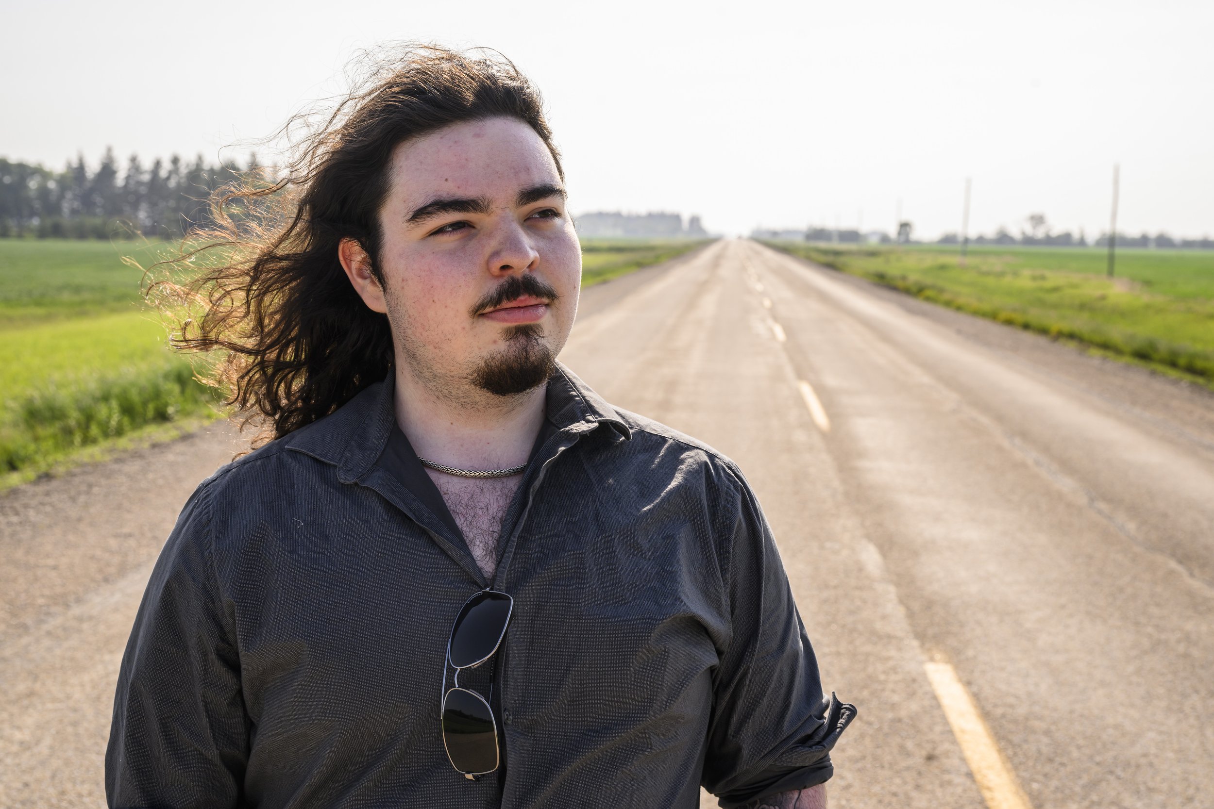 A man with long hair and a goatee standing on an empty rural road with green fields on either side during daytime.