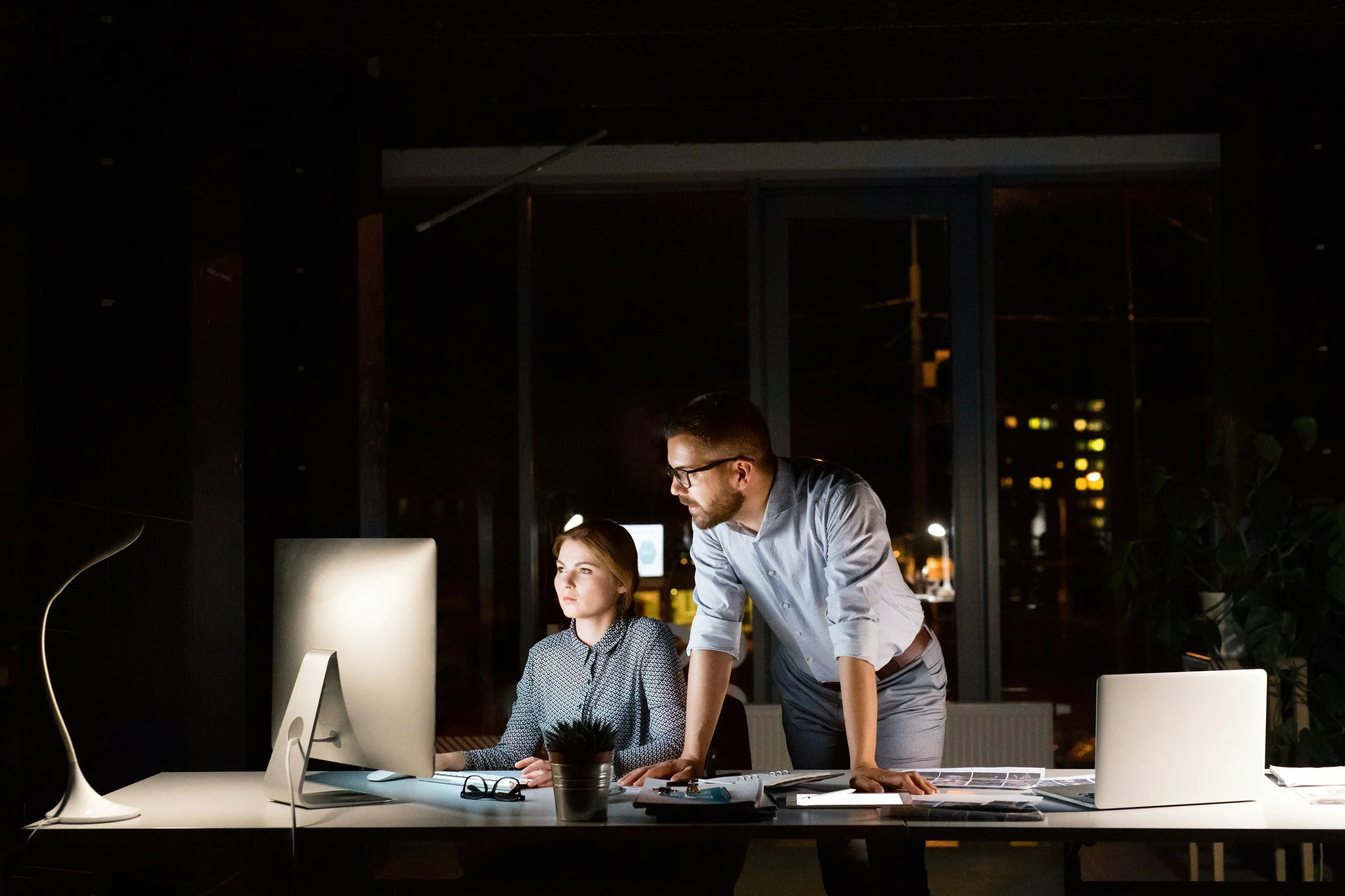 A man and a woman working together at a desk in an office at night, with the woman sitting at a computer and the man leaning over her, both focused on the screen.