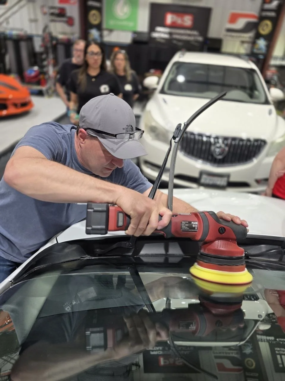 Man polishing car with electric buffer in auto repair shop, with three women and various cars and automotive equipment in background.