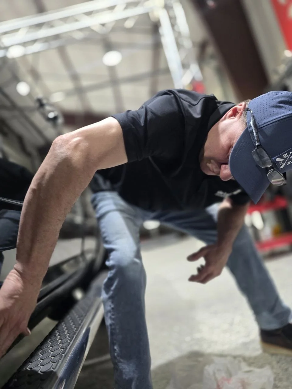 A man leaning forward with hands on a metal surface, wearing a navy baseball cap, glasses, a black shirt, and jeans, in an industrial or warehouse environment.