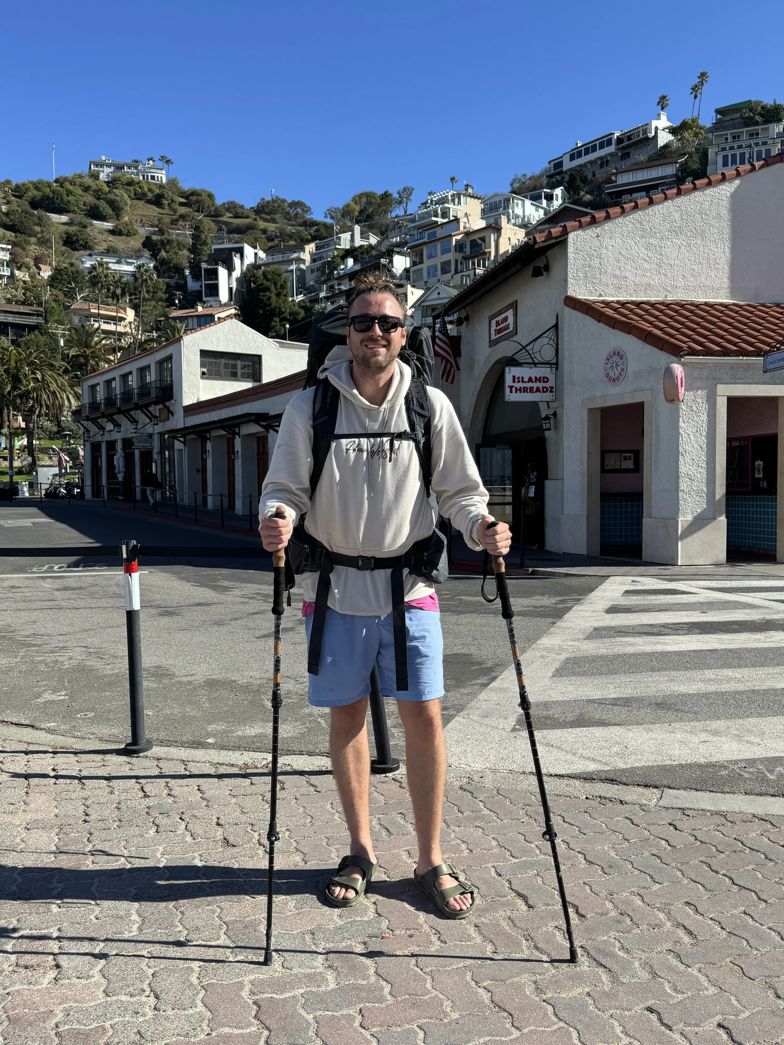 A young man wearing sunglasses, a beige hoodie, blue shorts, and sandals, standing on a city street with hiking poles and a backpack.