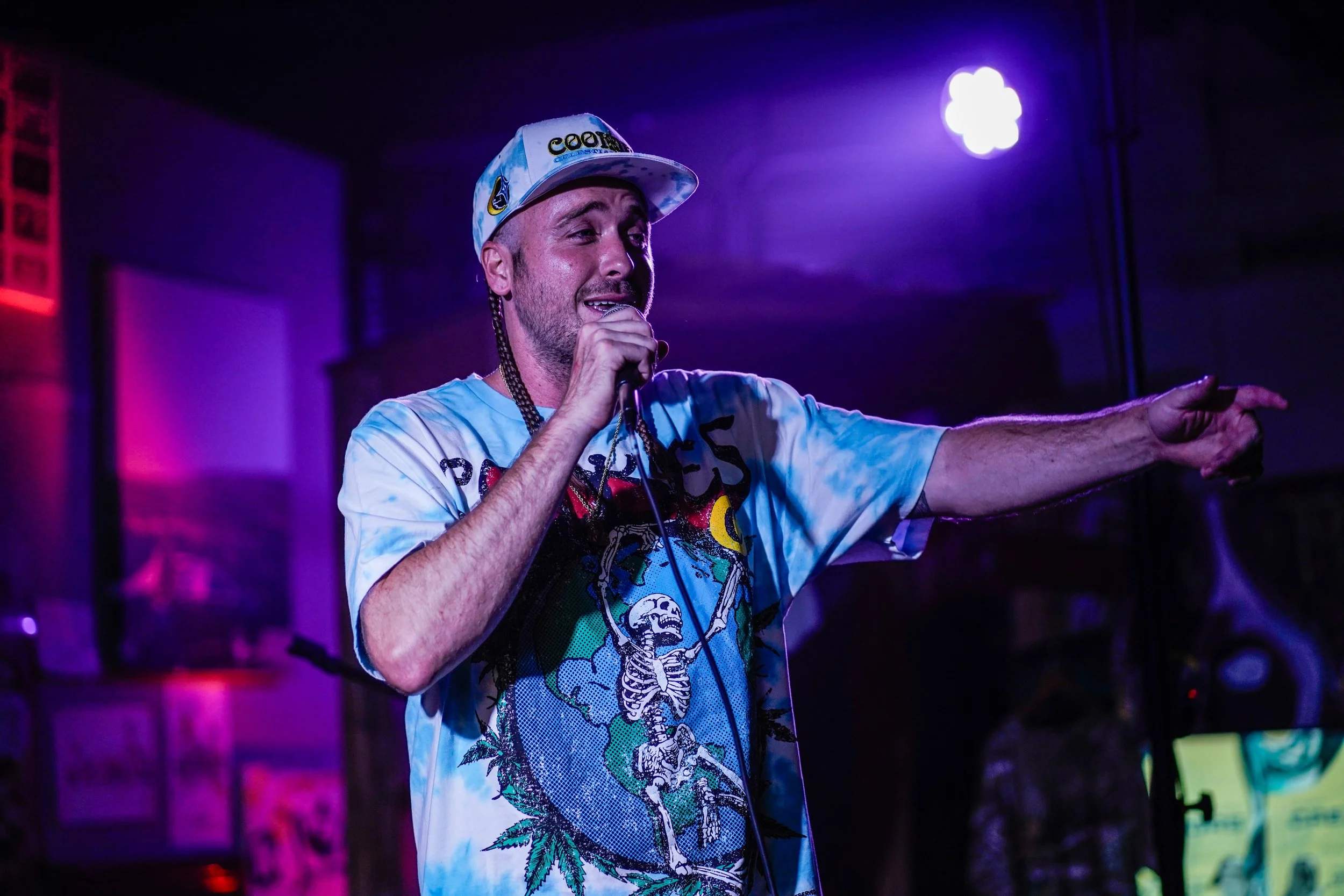 A man wearing a cap and a graphic t-shirt with a skeleton and cannabis leaf design, speaking into a microphone and pointing, with purple and blue stage lighting.