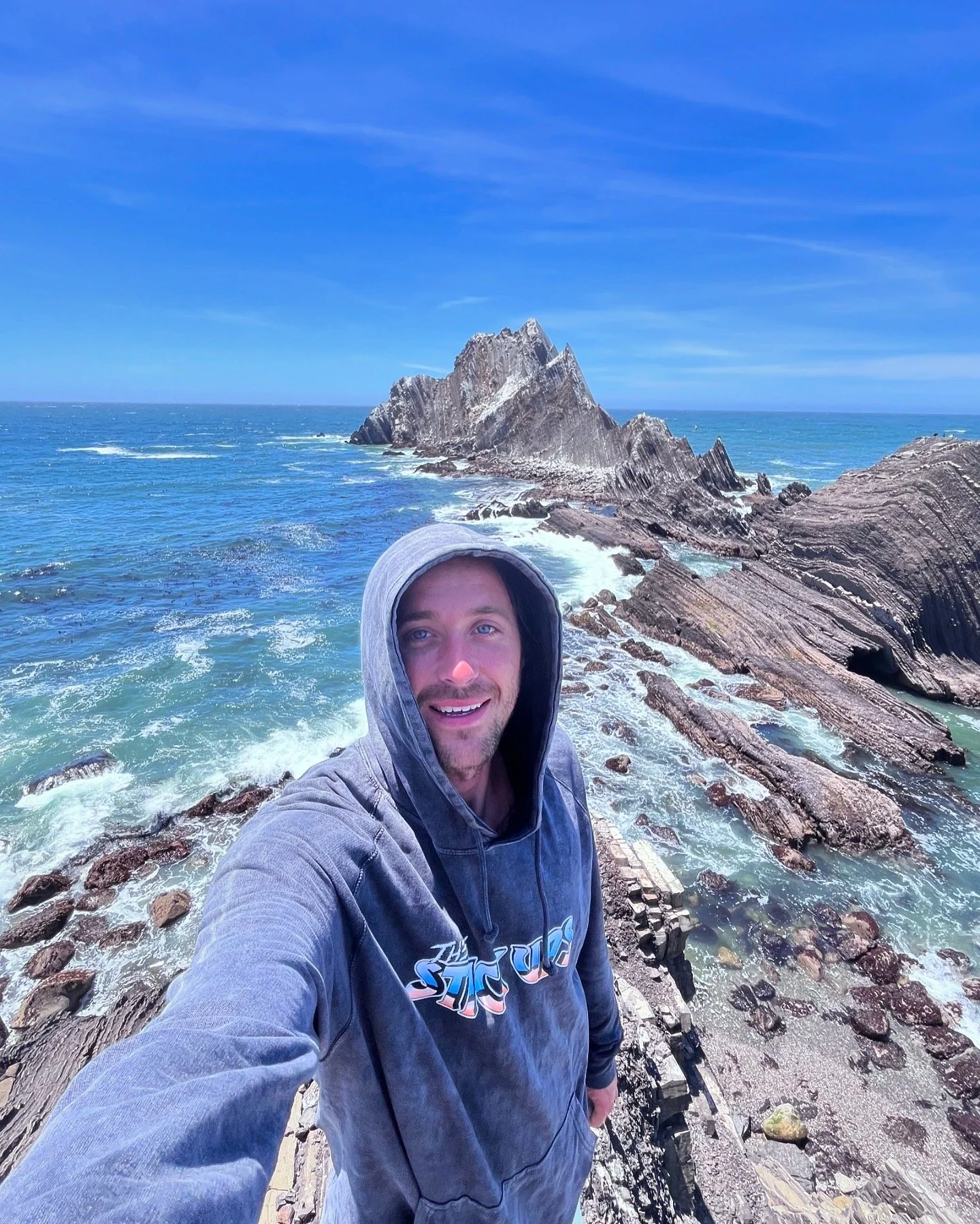 A man wearing a hoodie taking a selfie on a rocky coastline with blue ocean waves and a rocky island in the background under a clear blue sky.