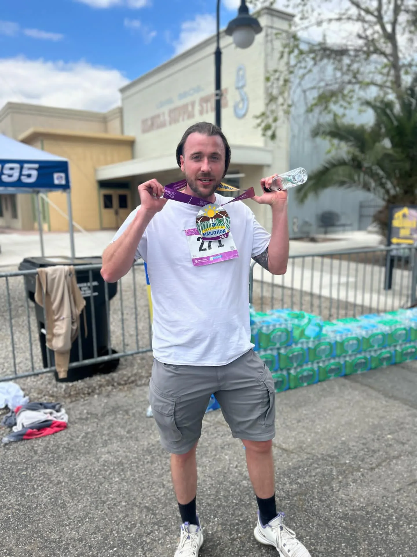 A man standing outside after completing a marathon, wearing a white t-shirt, gray shorts, and holding a water bottle and a medal around his neck. Behind him are water bottles and a building with a race-related sign.