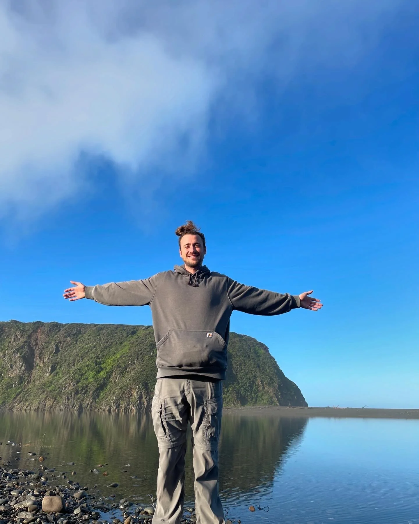 A smiling man stands on a rocky beach with arms outstretched, a body of water and a green mountainous landscape in the background under a blue sky with some clouds.