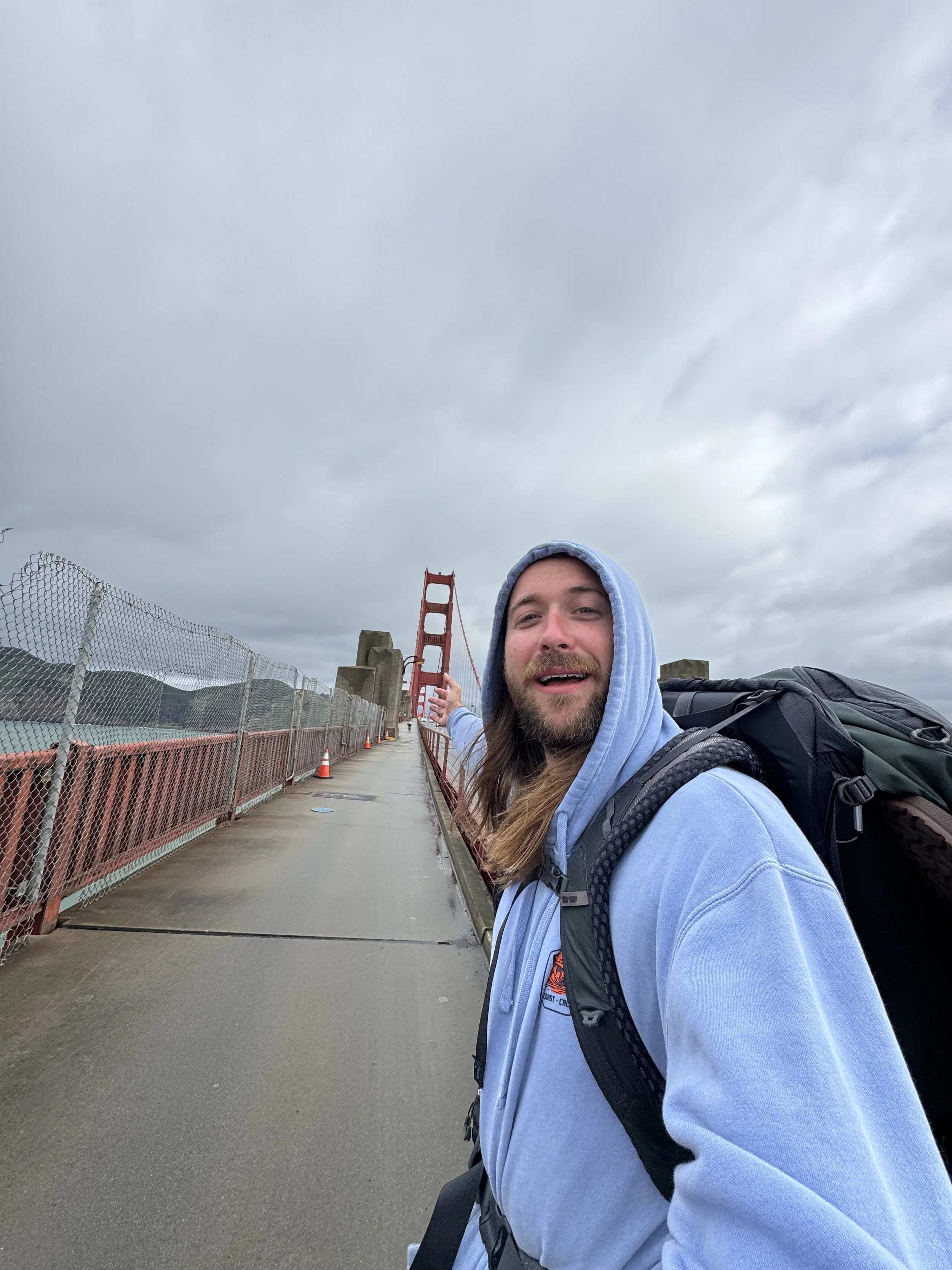 Young man with long hair and beard taking a selfie on the Golden Gate Bridge, pointing towards the bridge, wearing a blue hoodie and backpack on a cloudy day.