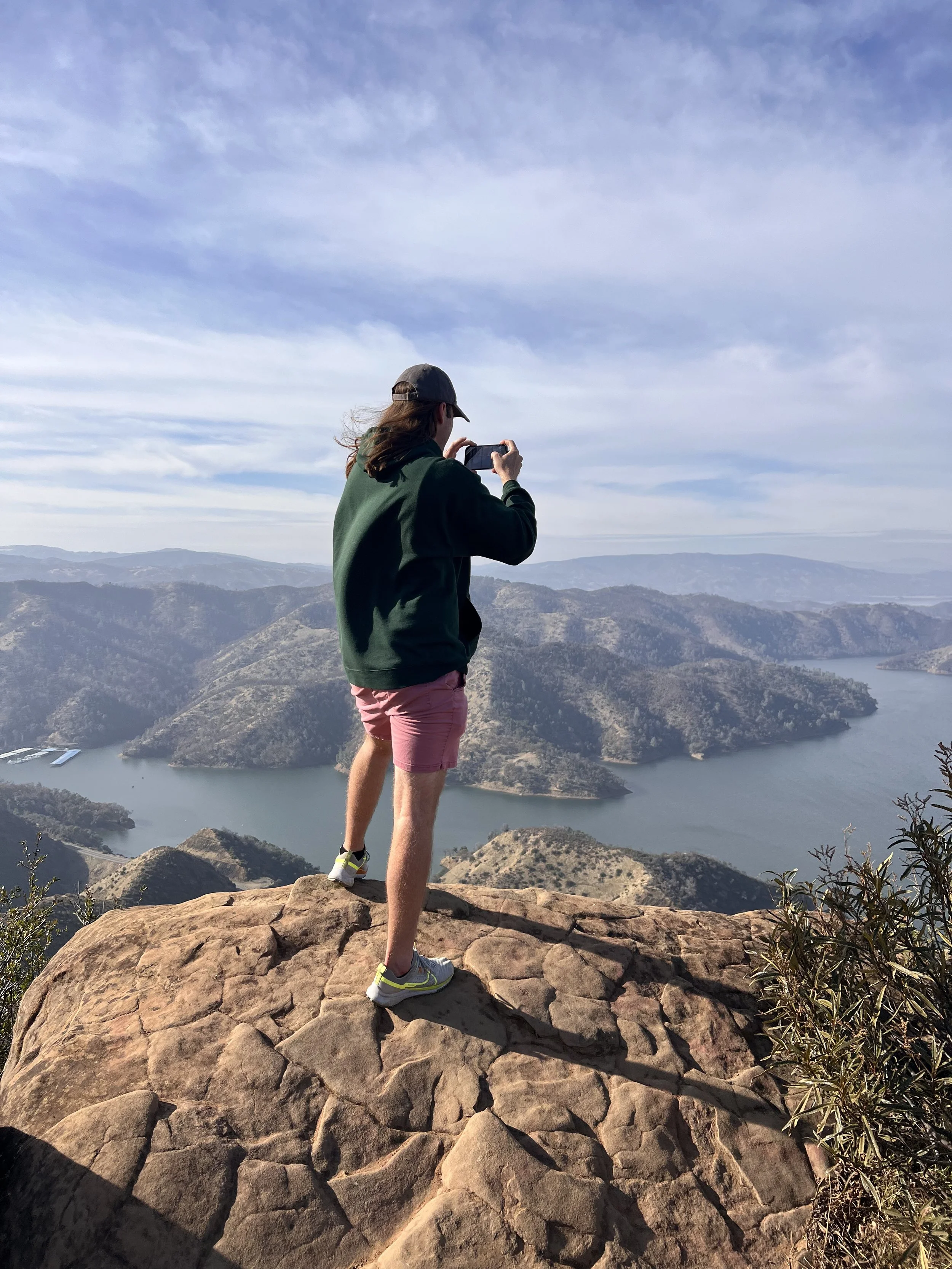 A person standing on a rocky ledge taking a photo with a smartphone of a scenic view of a lake and mountain landscape under a partly cloudy sky.