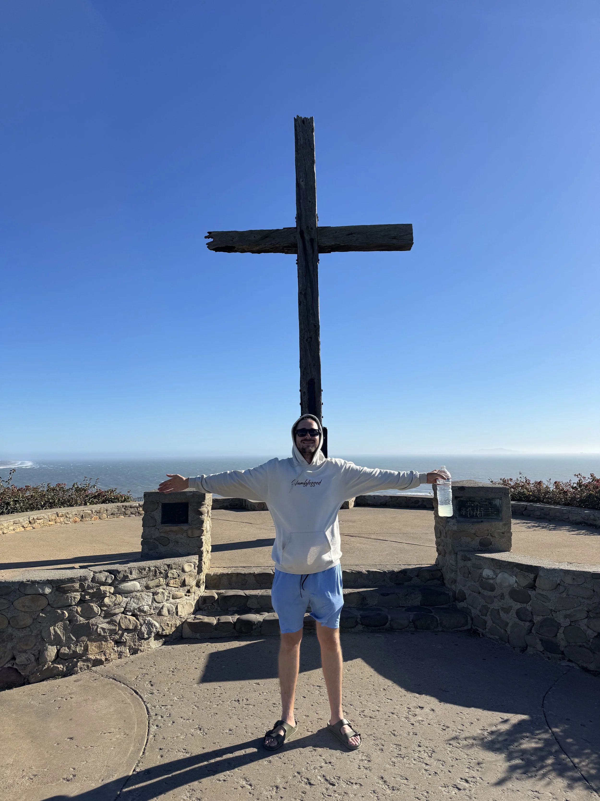 Man standing with arms outstretched at a scenic viewpoint with a large wooden cross and ocean in the background on a clear, sunny day.