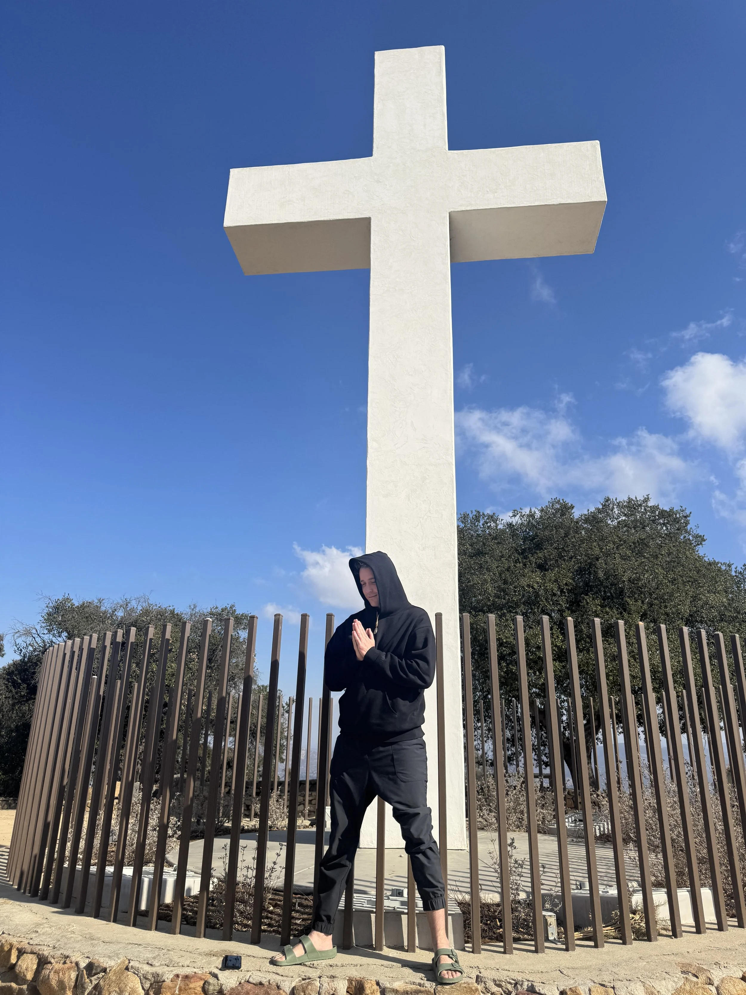 Person in black hoodie and pants with sandals standing in front of a large white cross monument on a bright, partly cloudy day.