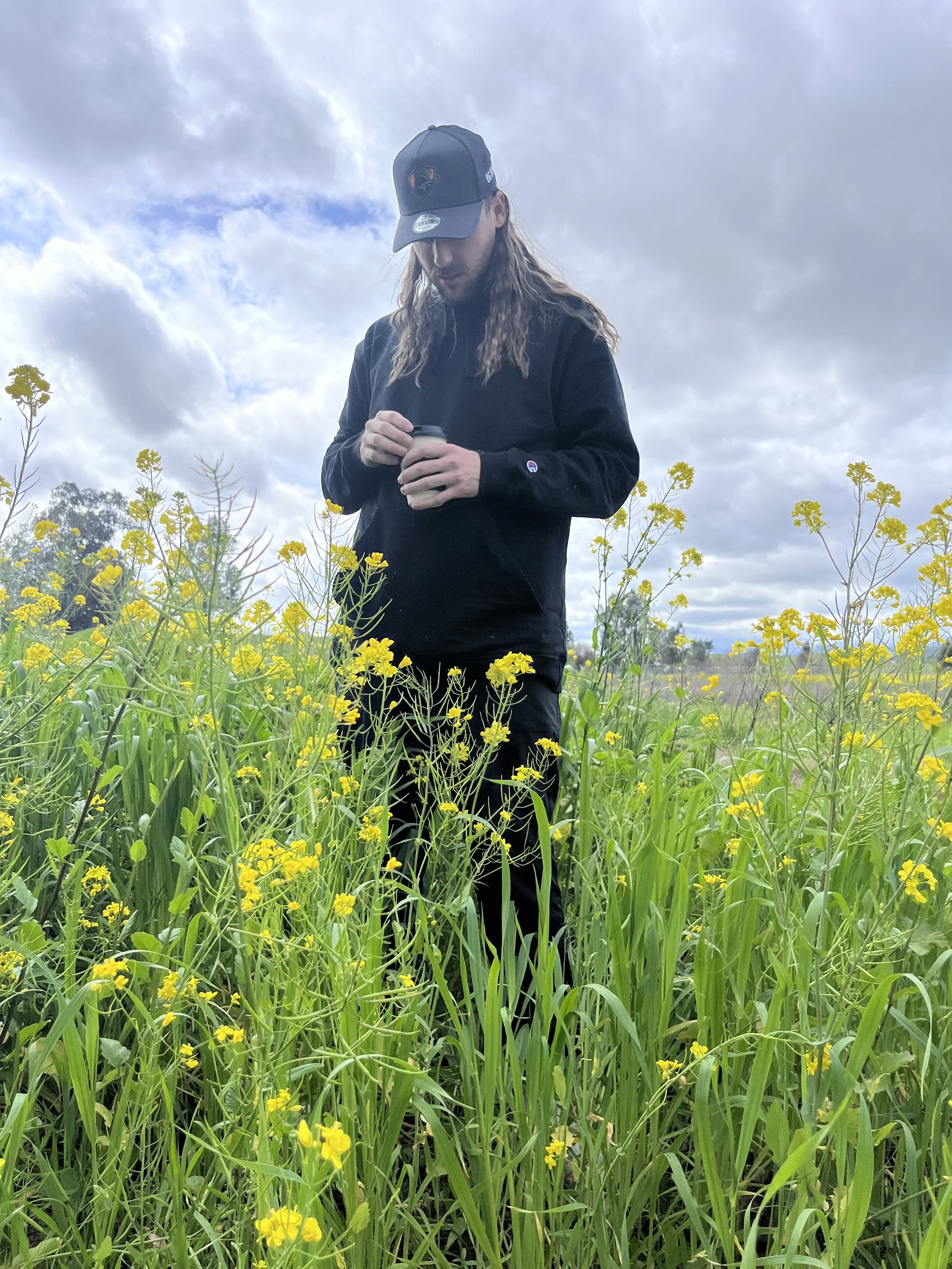 A man with long hair, wearing a black cap and sweatshirt, standing in a field of yellow flowers, holding a cup, with a cloudy sky in the background.