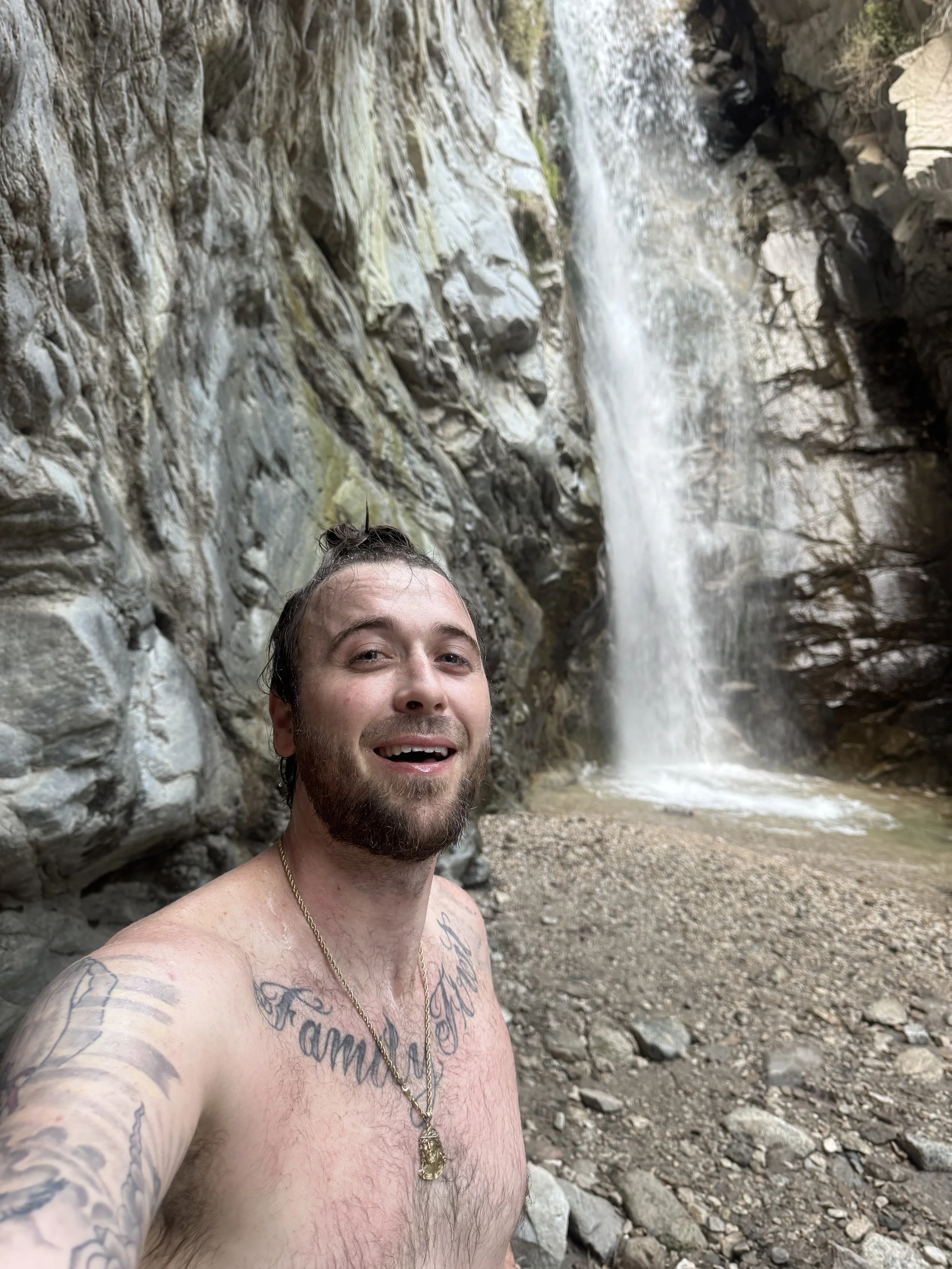 A man with tattoos and a gold chain taking a selfie in front of a waterfall in a rocky canyon.