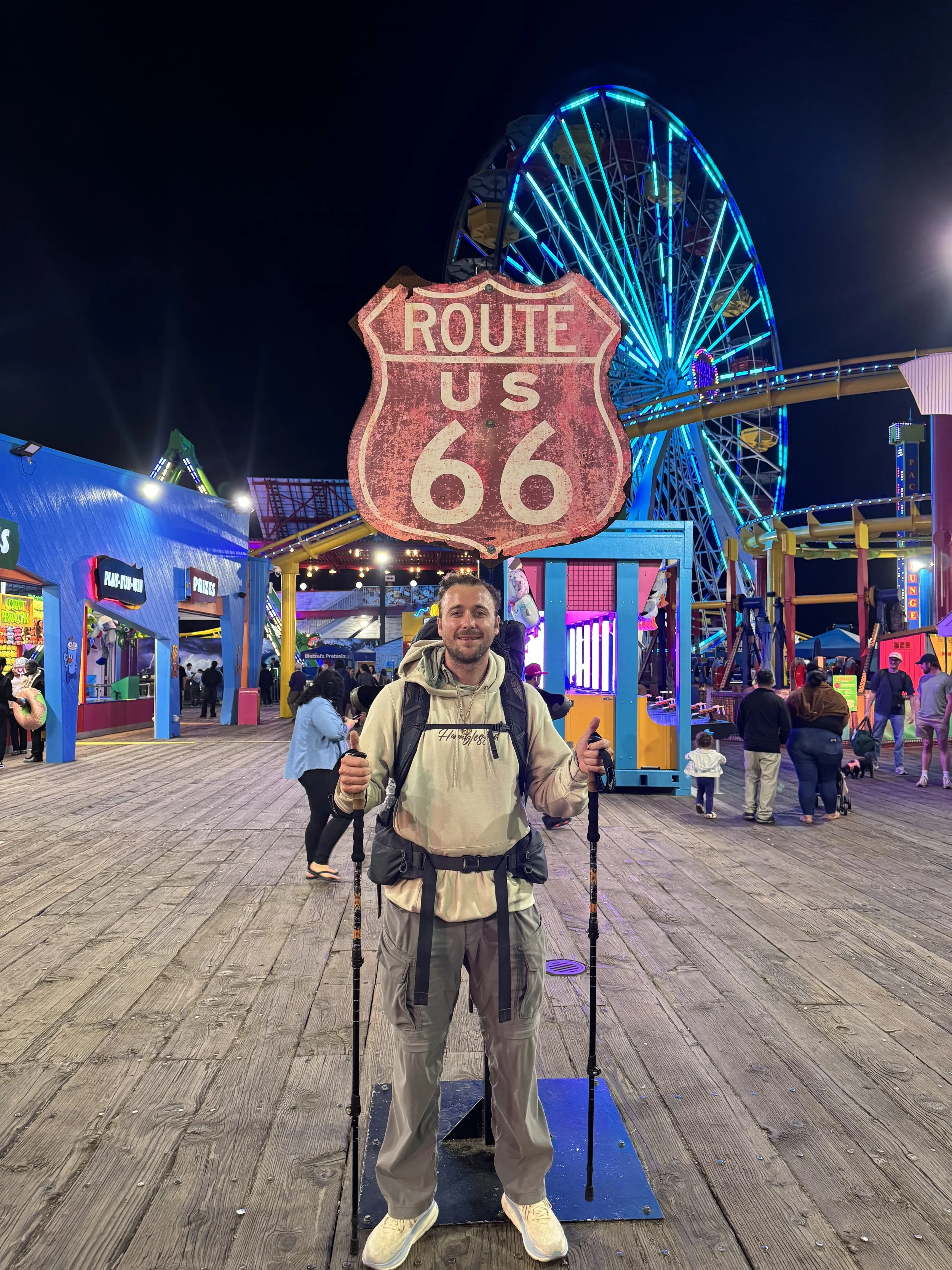 A man standing on a boardwalk at night, holding hiking poles, with a large sign reading 'Route U.S. 66' behind him. The amusement park rides and Ferris wheel are lit up with colorful lights in the background.