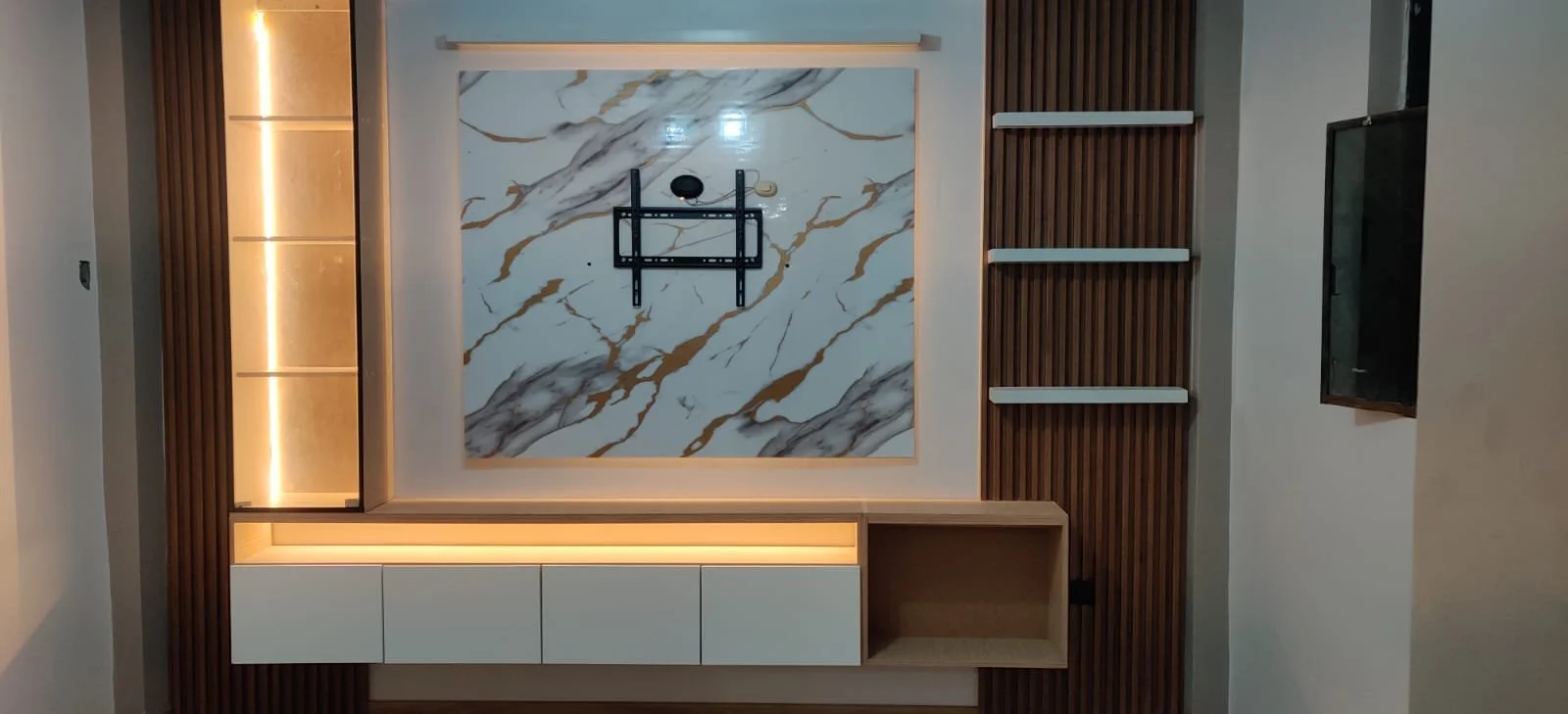 Decoración moderna en una pared con una mesa de centro de madera y estantes blancos en una pared con detalles de madera envejecida y un fondo de mármol con vetas doradas, luces blancas integradas y un televisor.