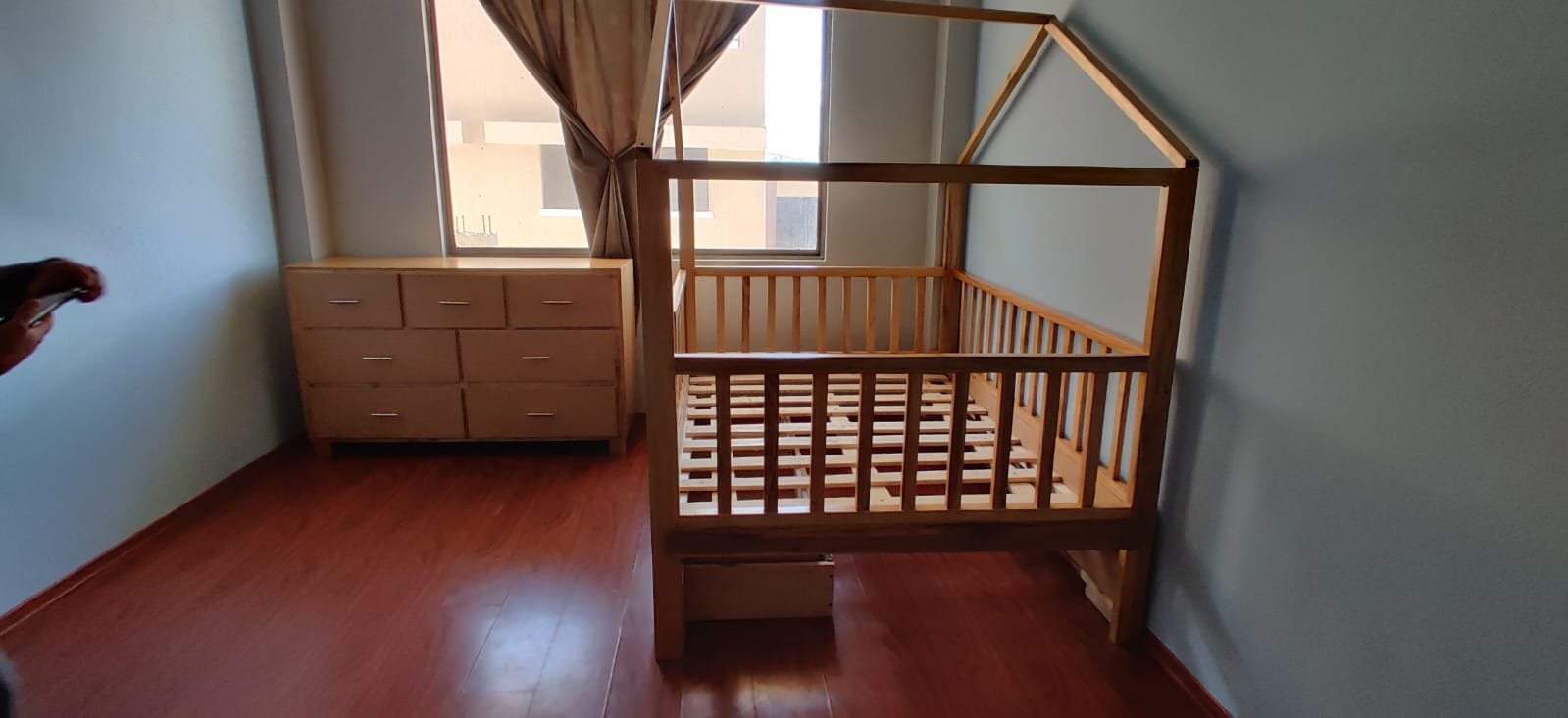 Cuna de madera sin colchón en una habitación con suelo de madera y una ventana con cortina beige, y un mueble de madera con varios cajones.