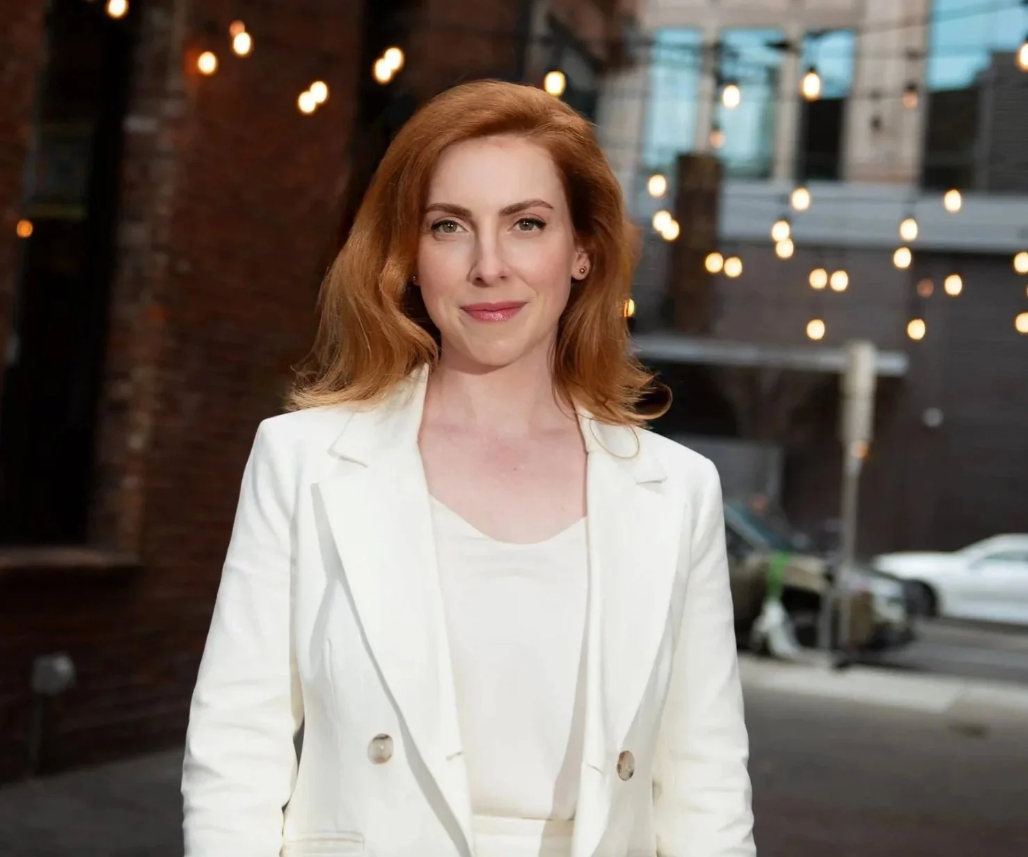 A woman with red hair wearing a white blazer outdoors with string lights and buildings in the background.