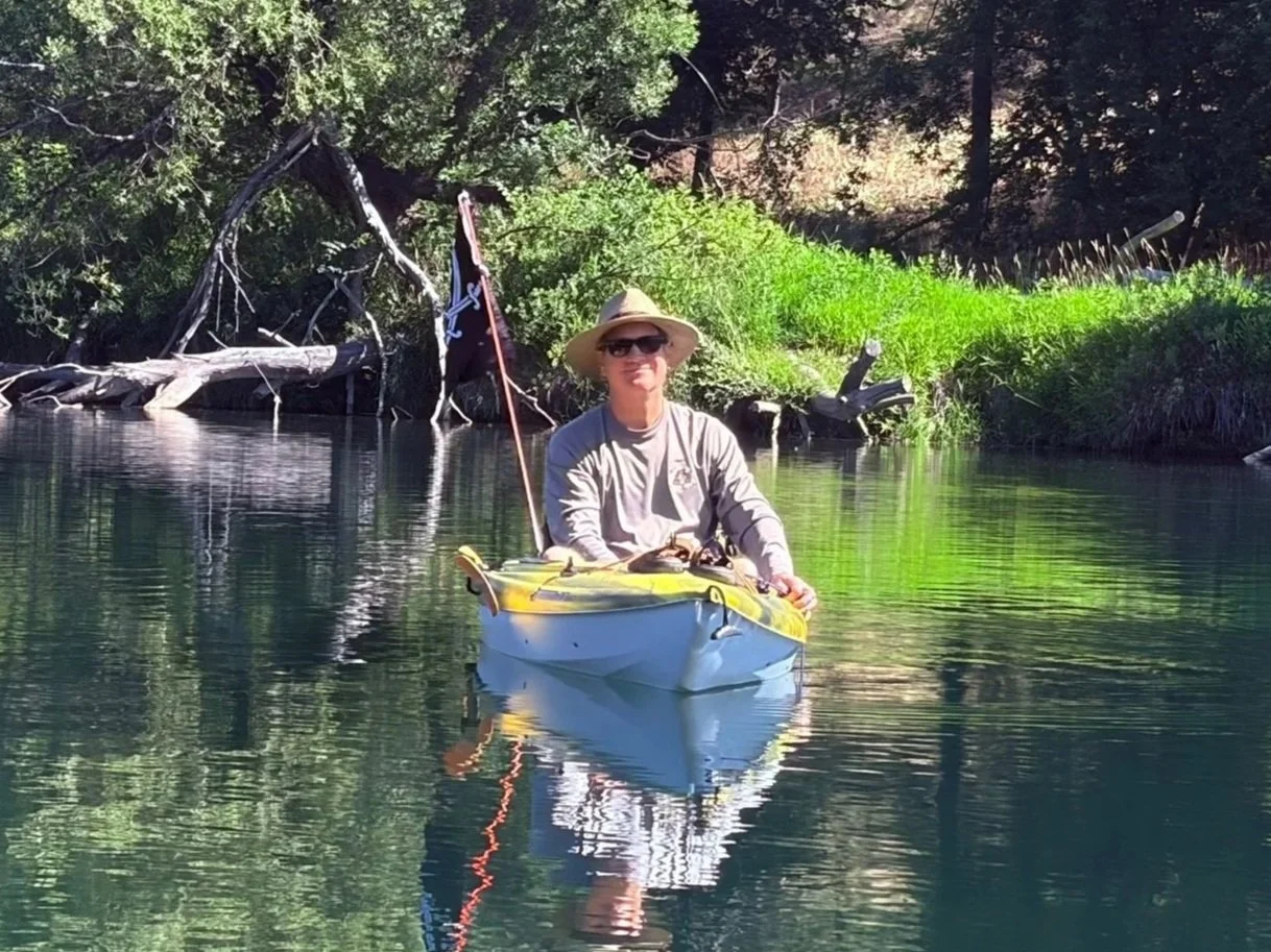 A person wearing a wide-brimmed hat and sunglasses sits in a yellow and blue kayak on a calm river, with green trees and fallen logs in the background.