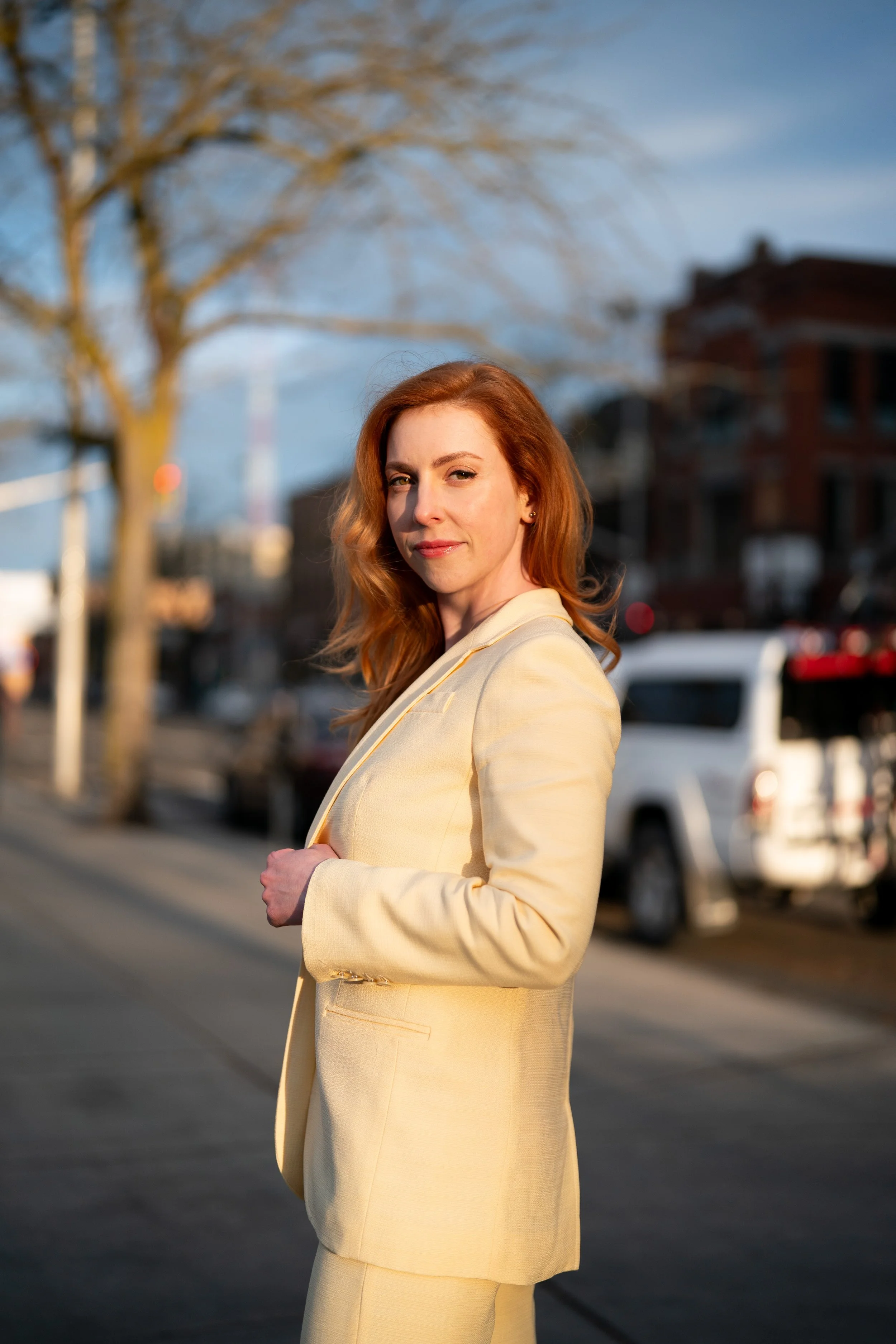 A woman with red hair wearing a dark, cream-colored suit standing outdoors on a street during sunset, with cars and buildings in the background.