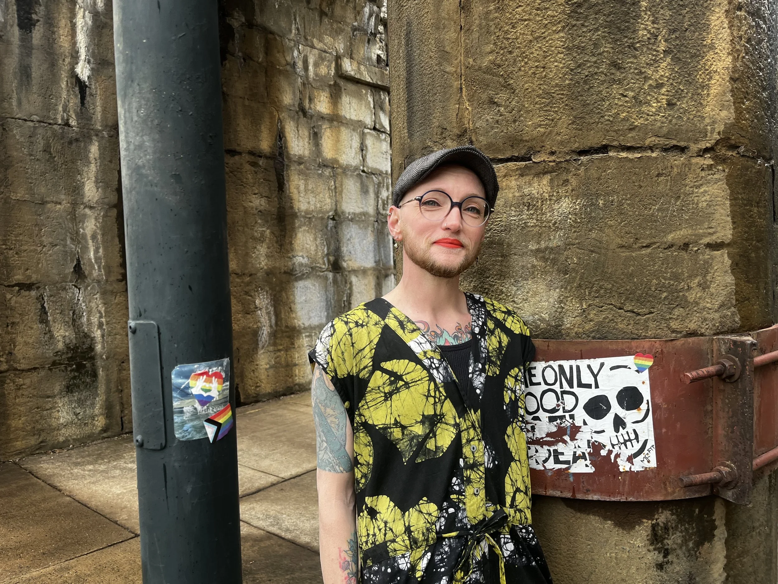 Jordan Zipser, a white nonbinary person with a beard, smiles slightly at the camera. they look head-on while wearing a grey flat cap, glasses, orangey lipstick, and a black, lime green, and white top. they have several visible tattoos.