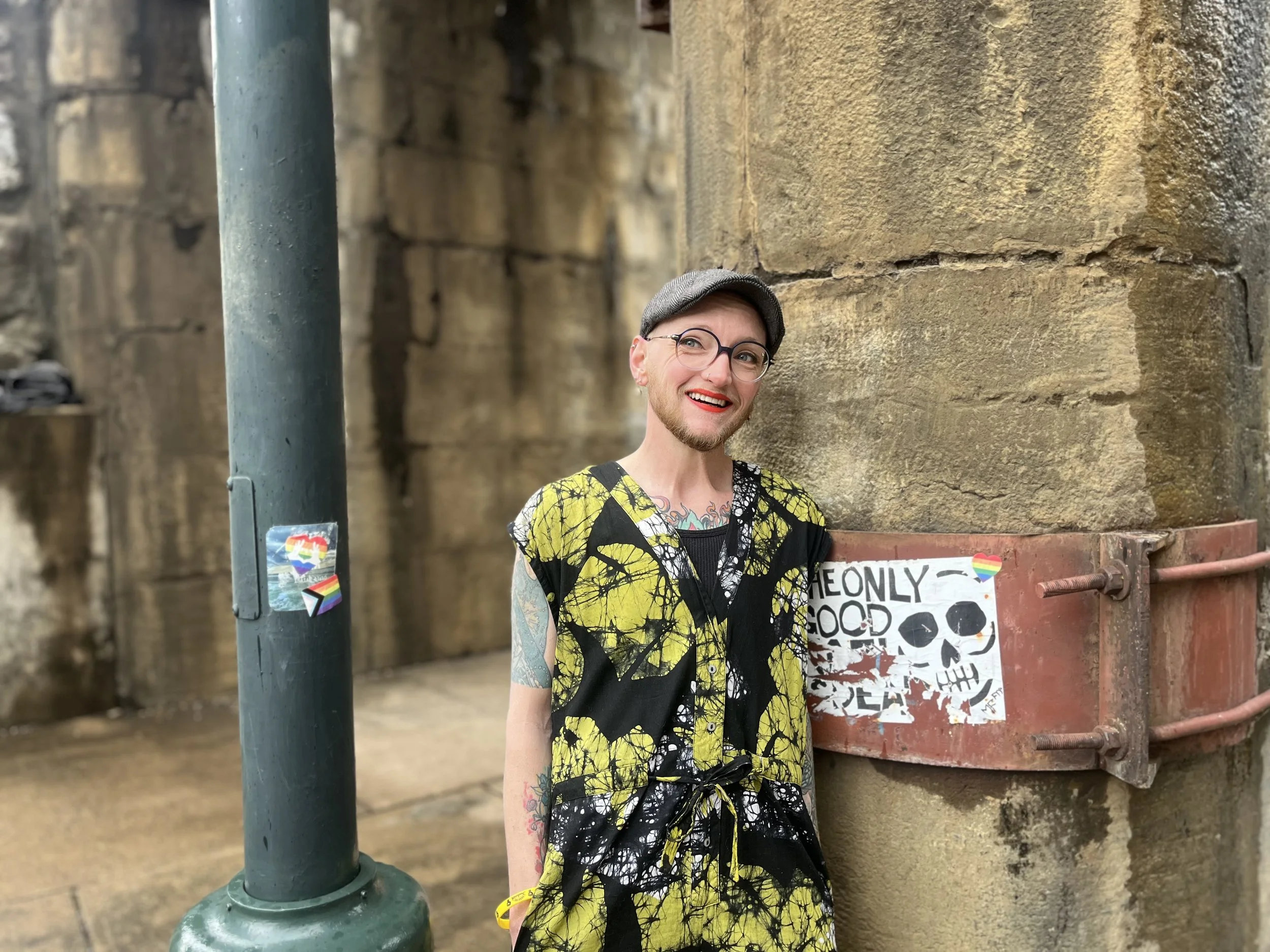 Jordan Zipser, a white nonbinary person, looks away from the camera and smiles. they wear glasses, a beard, a grey flat cap, orange lipstick, and a fun black, lime-green, and white patterned jumper.