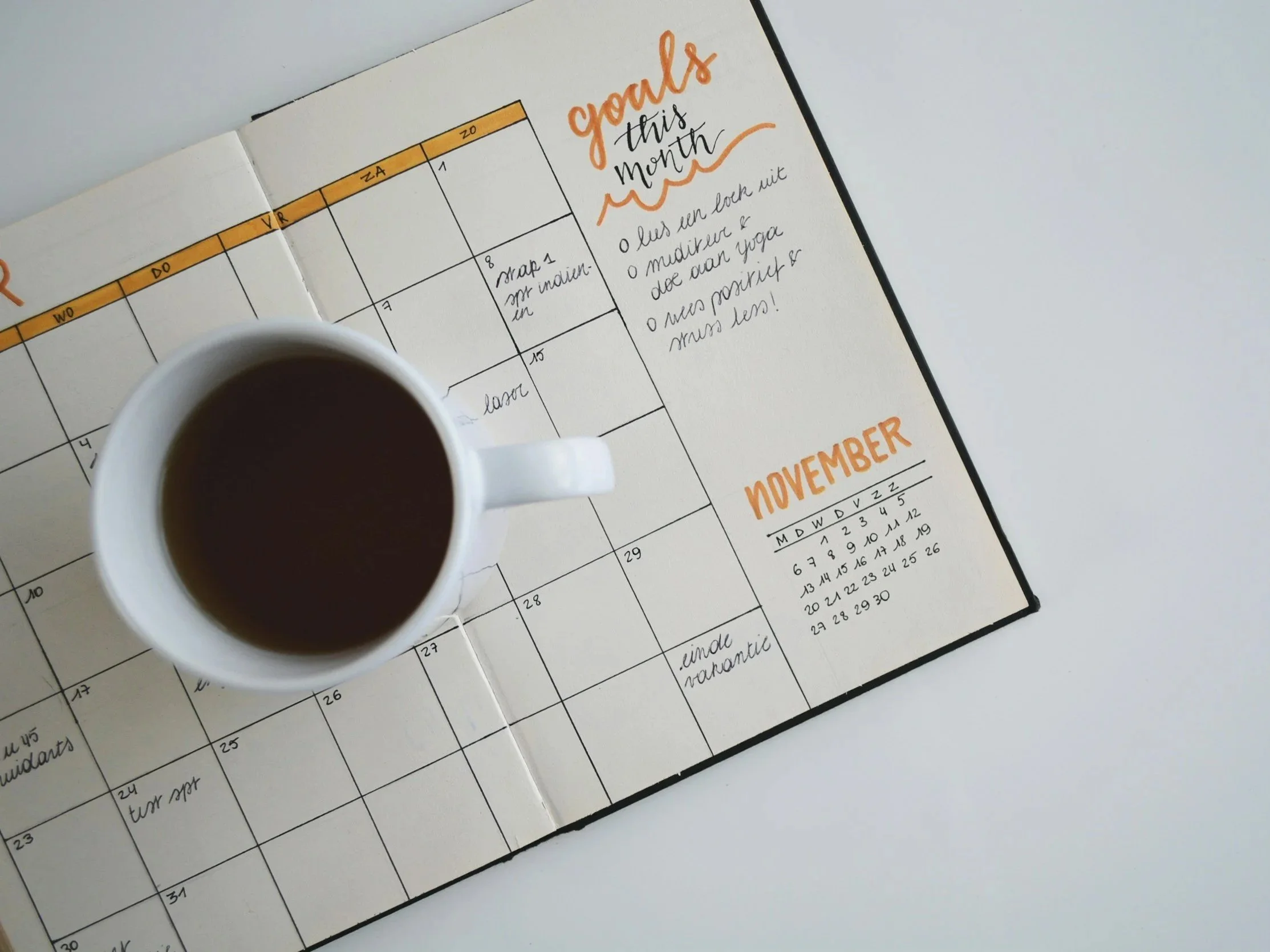 A calendar page for the month of November is open on a white surface, with handwritten notes and a colorful header. A cup of coffee is placed in front of the calendar, partially blocking it.