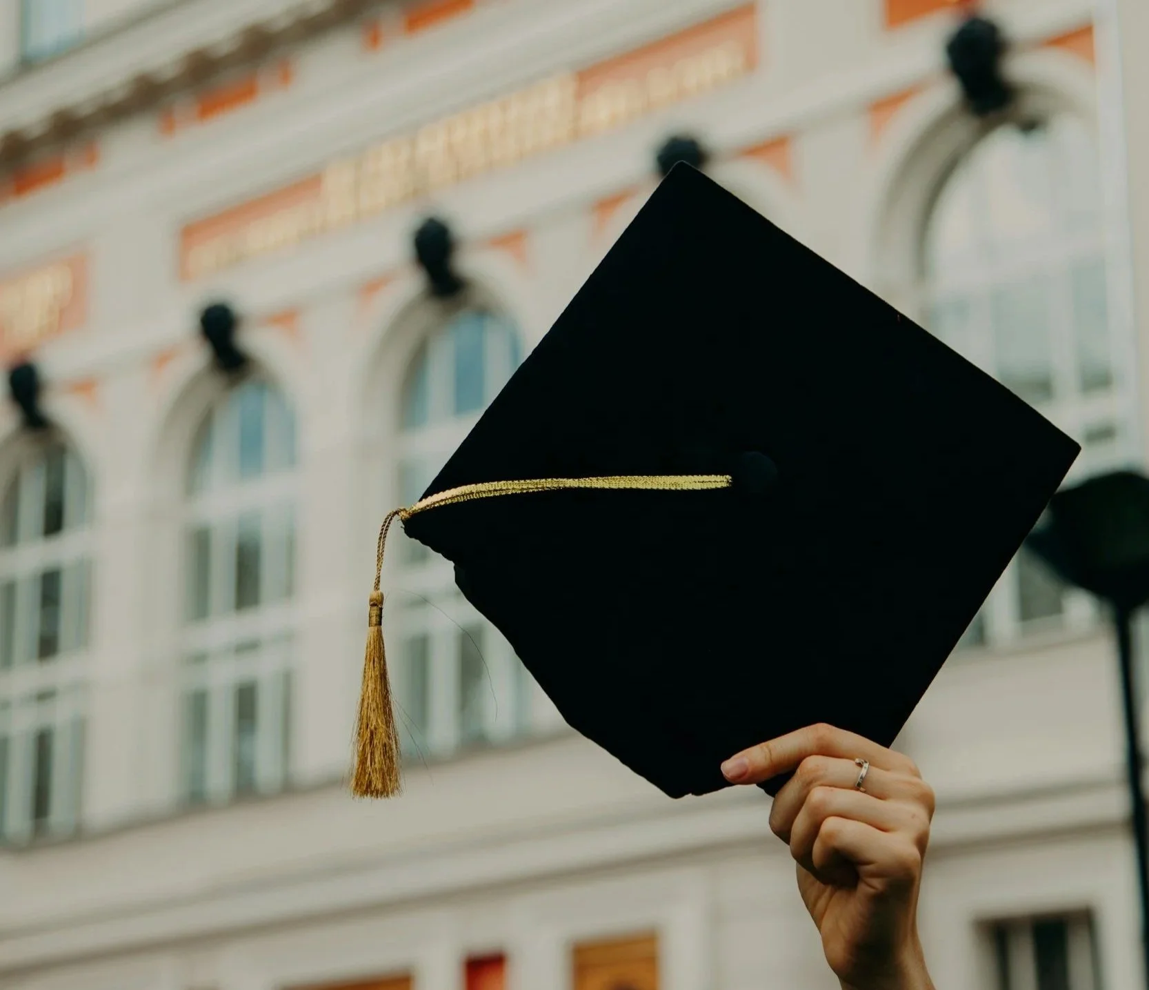 A person holding up a black graduation cap with a gold tassel, standing in front of a building with large arched windows.