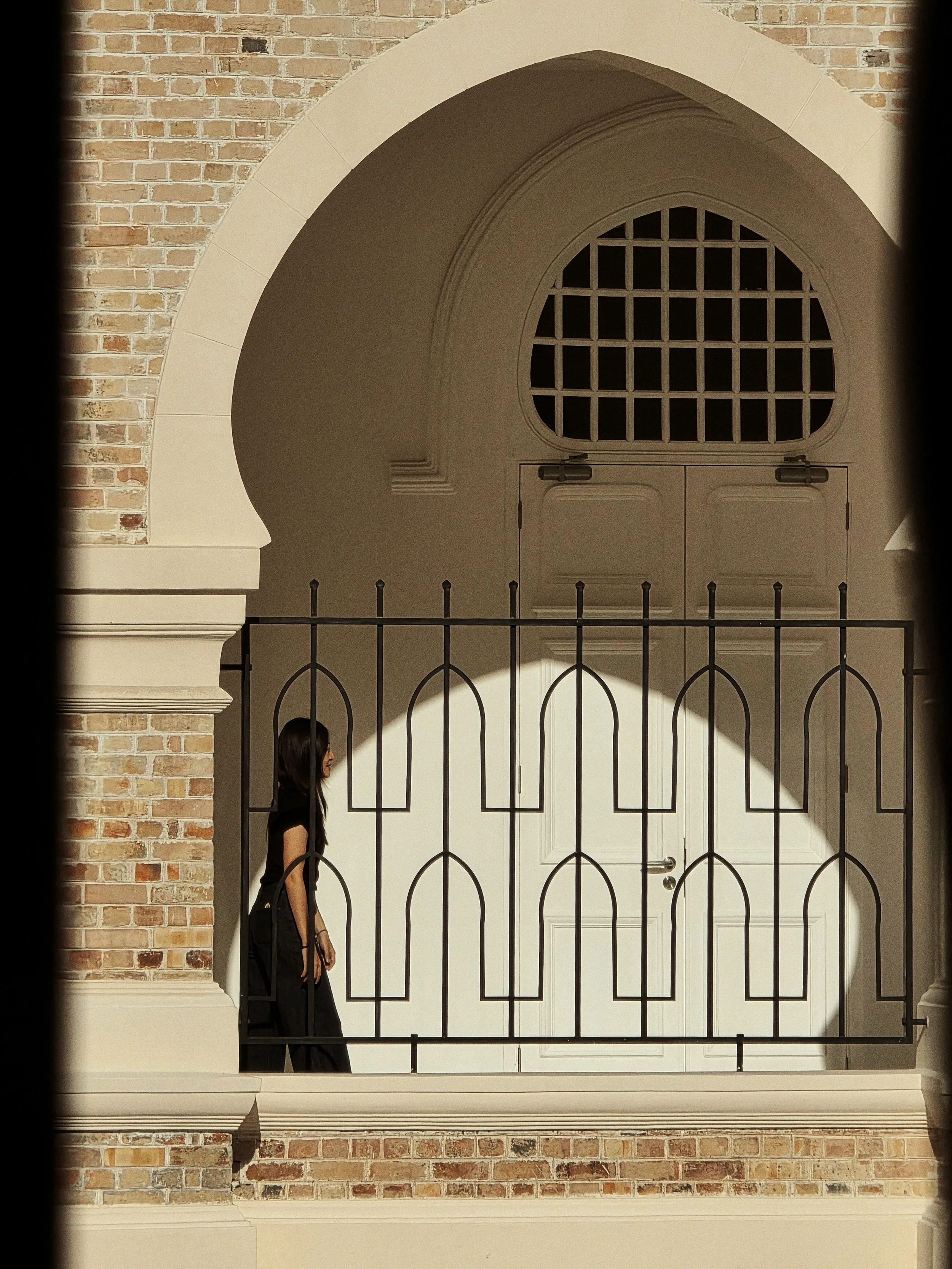 A woman with long black hair walking past a decorative black metal fence in front of a building with a brick exterior and arched white door and window.