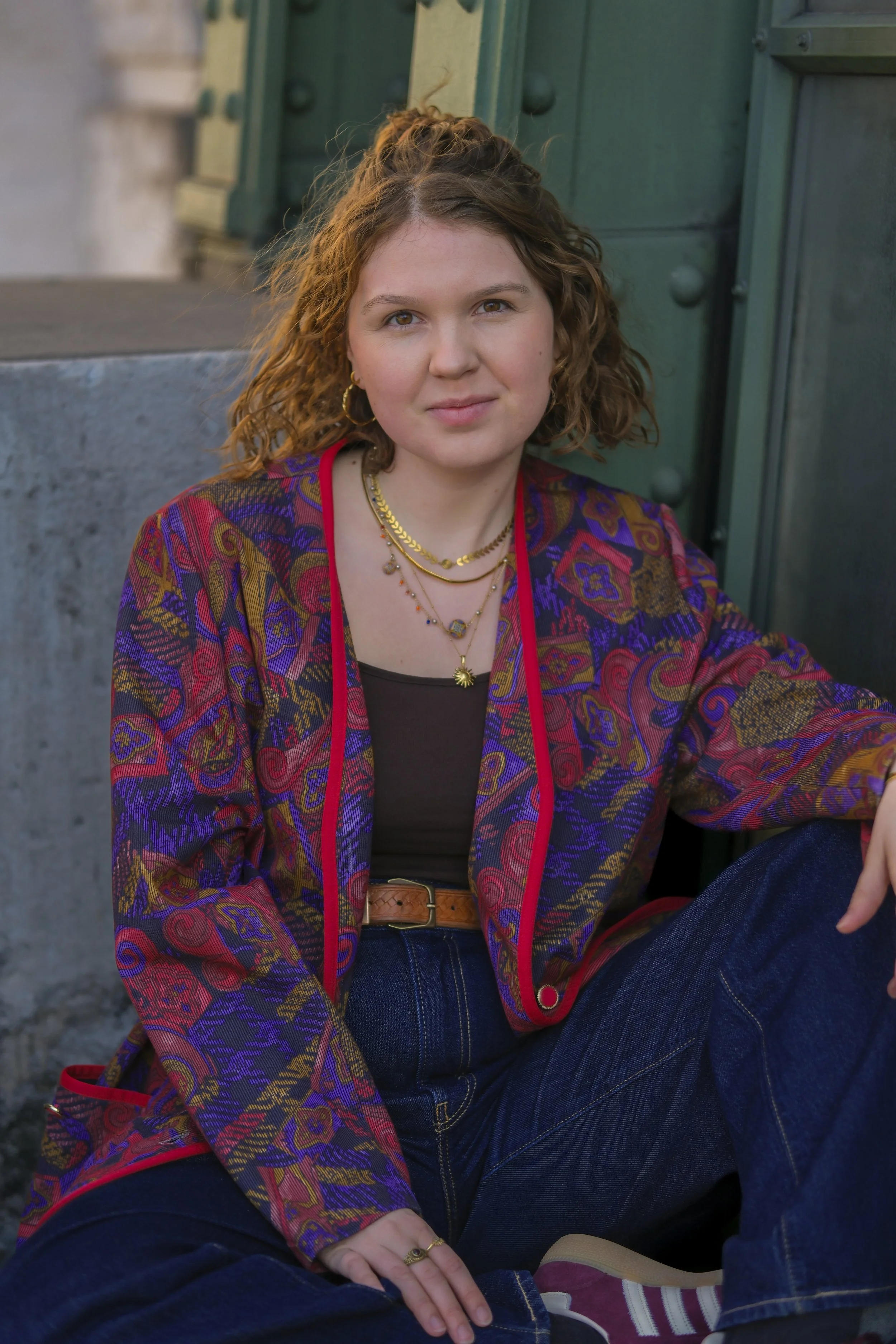 Eine junge Frau mit lockigem braunem Haar, die bunte Jacke mit roten, blauen und goldenen Mustern trägt, sitzt vor einer grünen Tür und schaut freundlich in die Kamera. Sie trägt goldene Ohrringe und mehrere Halsketten, sitzt entspannt auf einem Sitz."