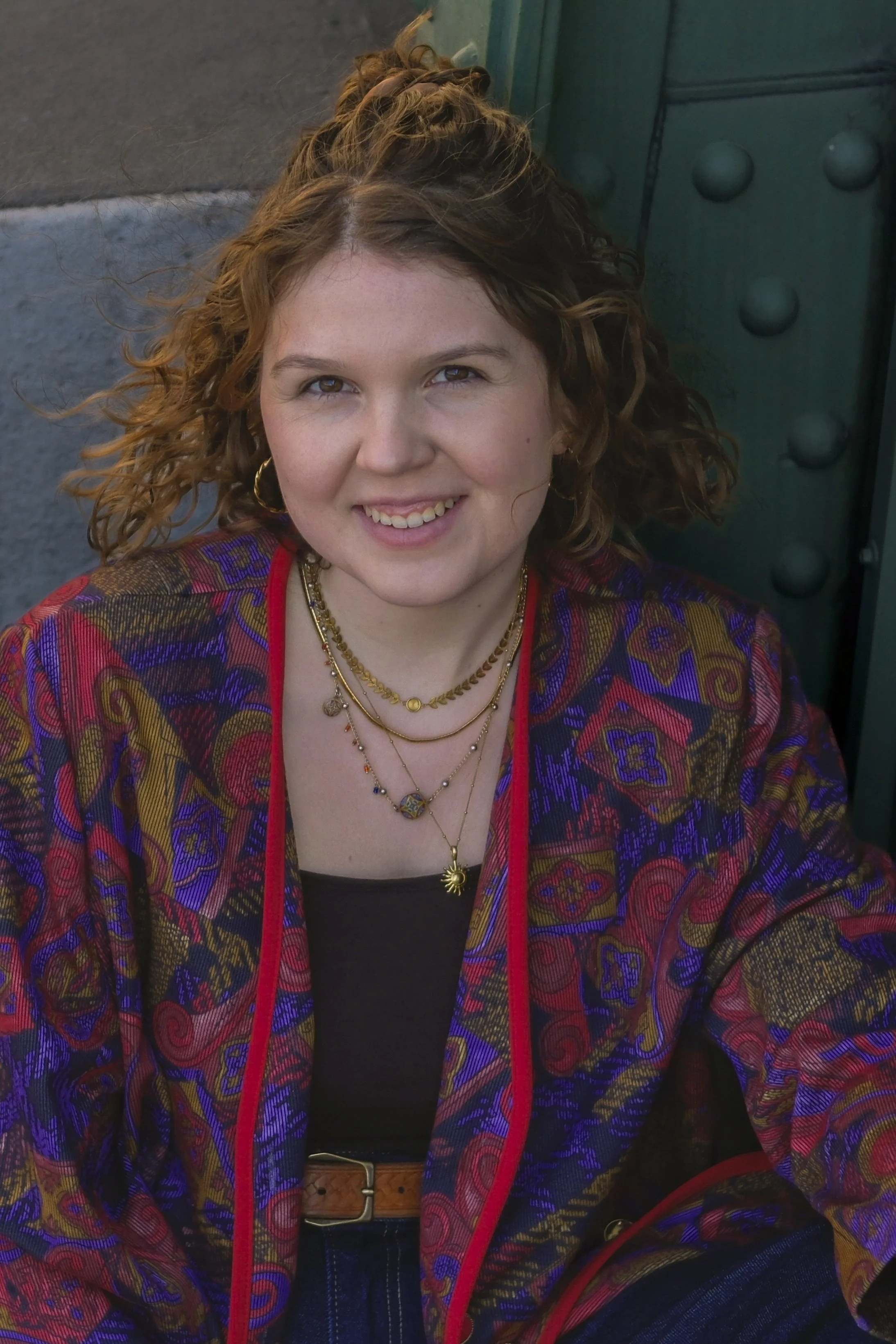 Junge Frau mit lockigem Haar, trägt ein bunter Jacke mit rotem und goldfarbenem Design, mehrere Halsketten, lächelt und sitzt an einem Tisch im Freien.