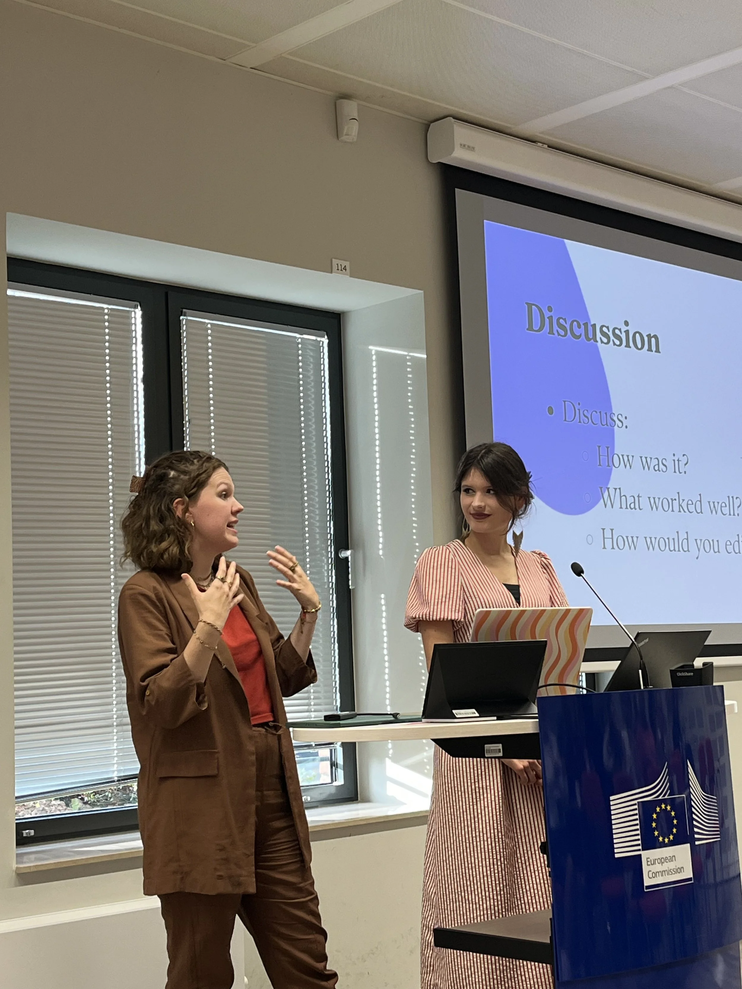Zwei Frauen sprechen während einer Präsentation im Konferenzraum der Europäischen Kommission. Im Hintergrund ist ein Projektorbild mit dem Wort 'Discussion' und Fragen sichtbar.