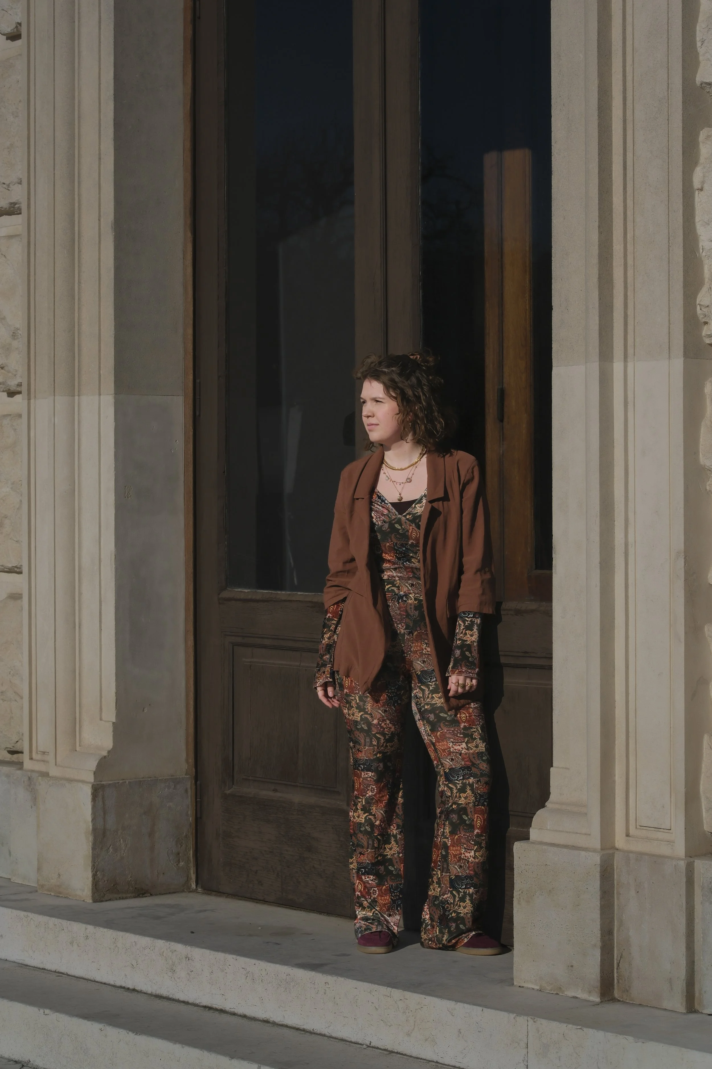Junge Frau steht vor einer alten Steintür, trägt eine braune Jacke und bunte, breit gemusterte Hose, schaut nach rechts, trägt mehrere Ketten, Sonnenlicht, Schatten, dunkelblauer Himmel im Hintergrund.