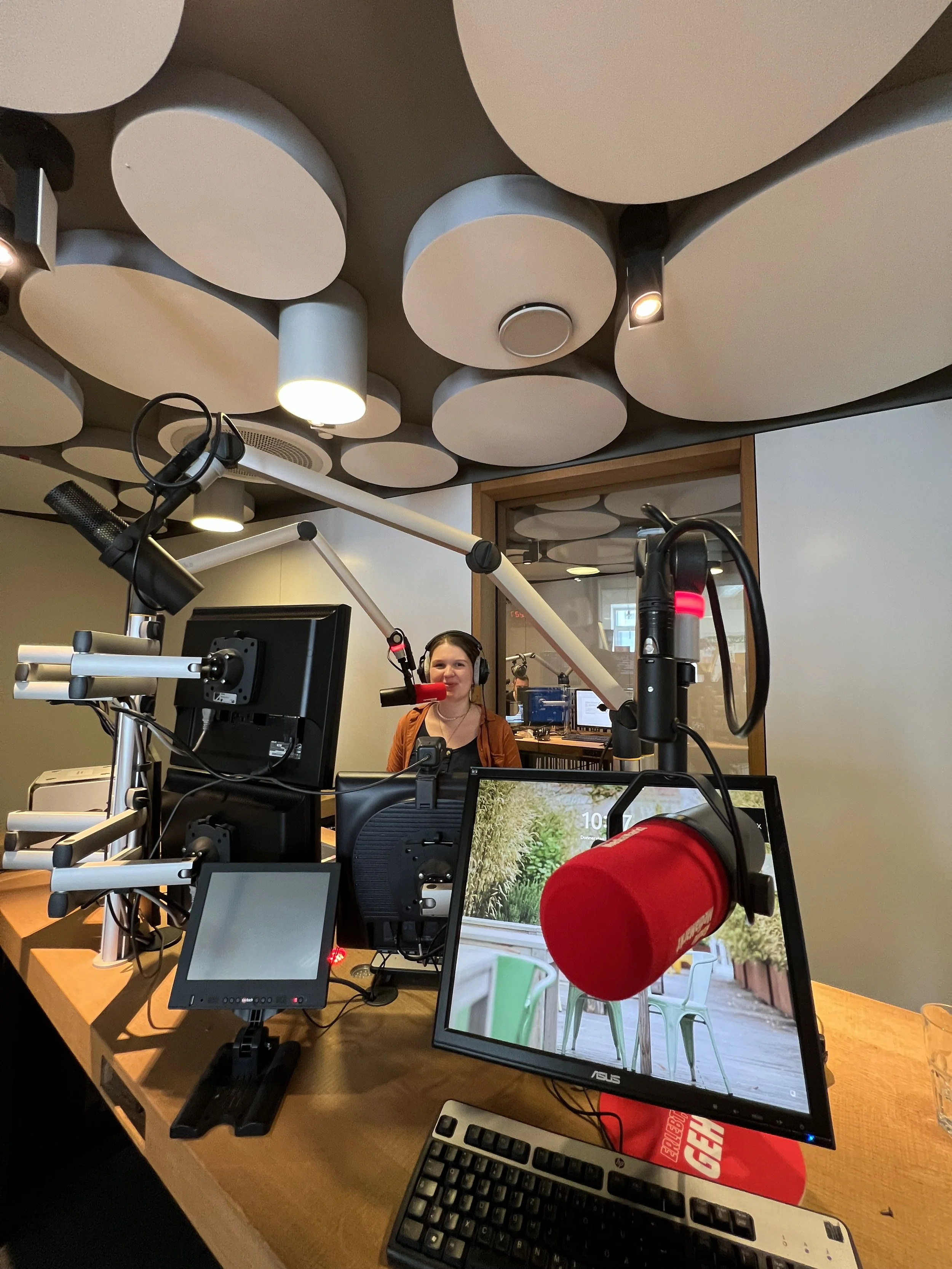 Aufnahme eines Aufnahmestudio mit Mikrofonen, Bildschirmen und einer Person, die im Hintergrund an dem Radiomikrofon steht. 