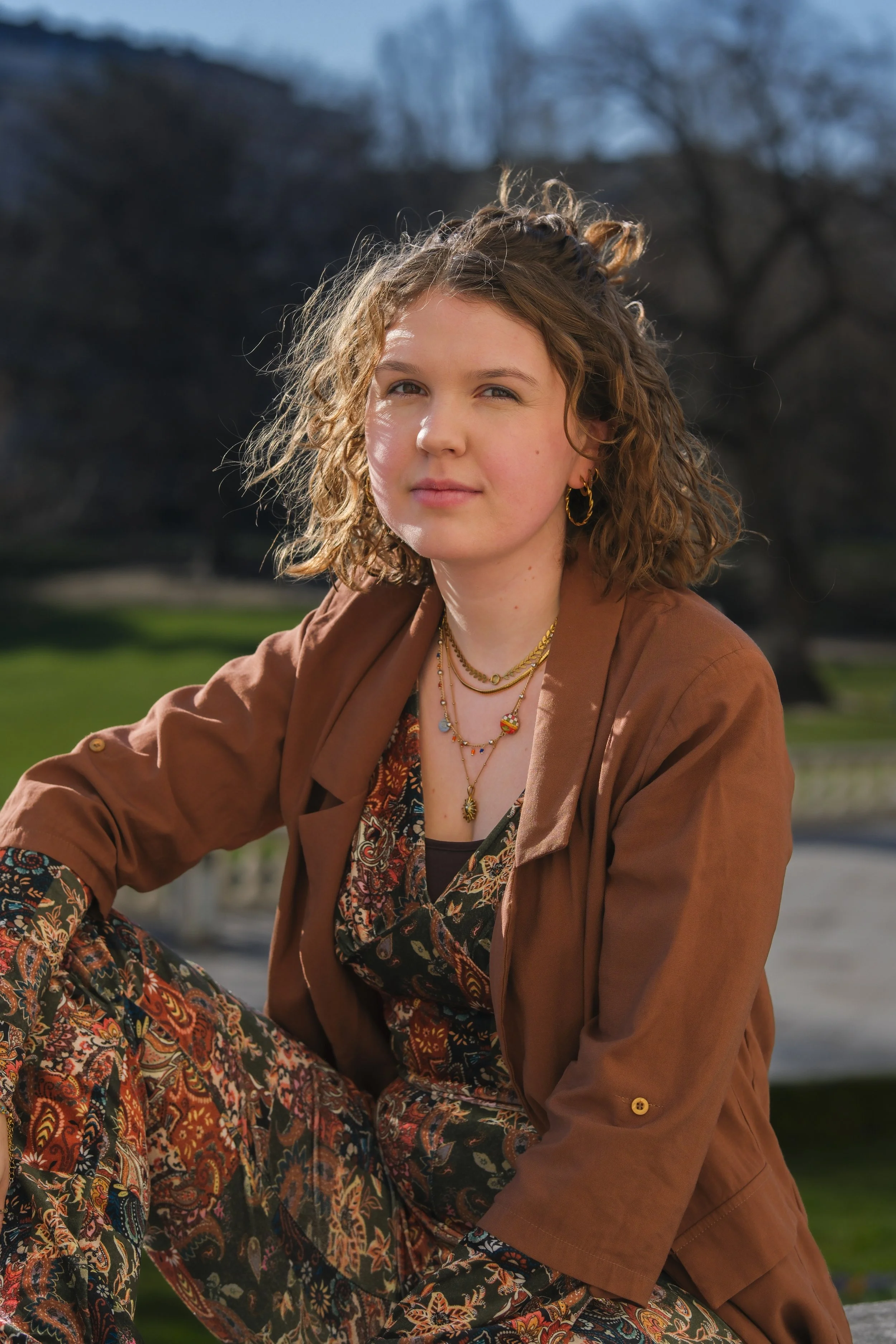 Junge Frau mit lockigem Haar, goldenen Ohrringen und mehreren Halsketten, sitzend im Freien bei Sonnenlicht, trägt ein braunes Jackett und ein buntes Kleid mit Paisleymuster, im Hintergrund Bäume und Himmel.