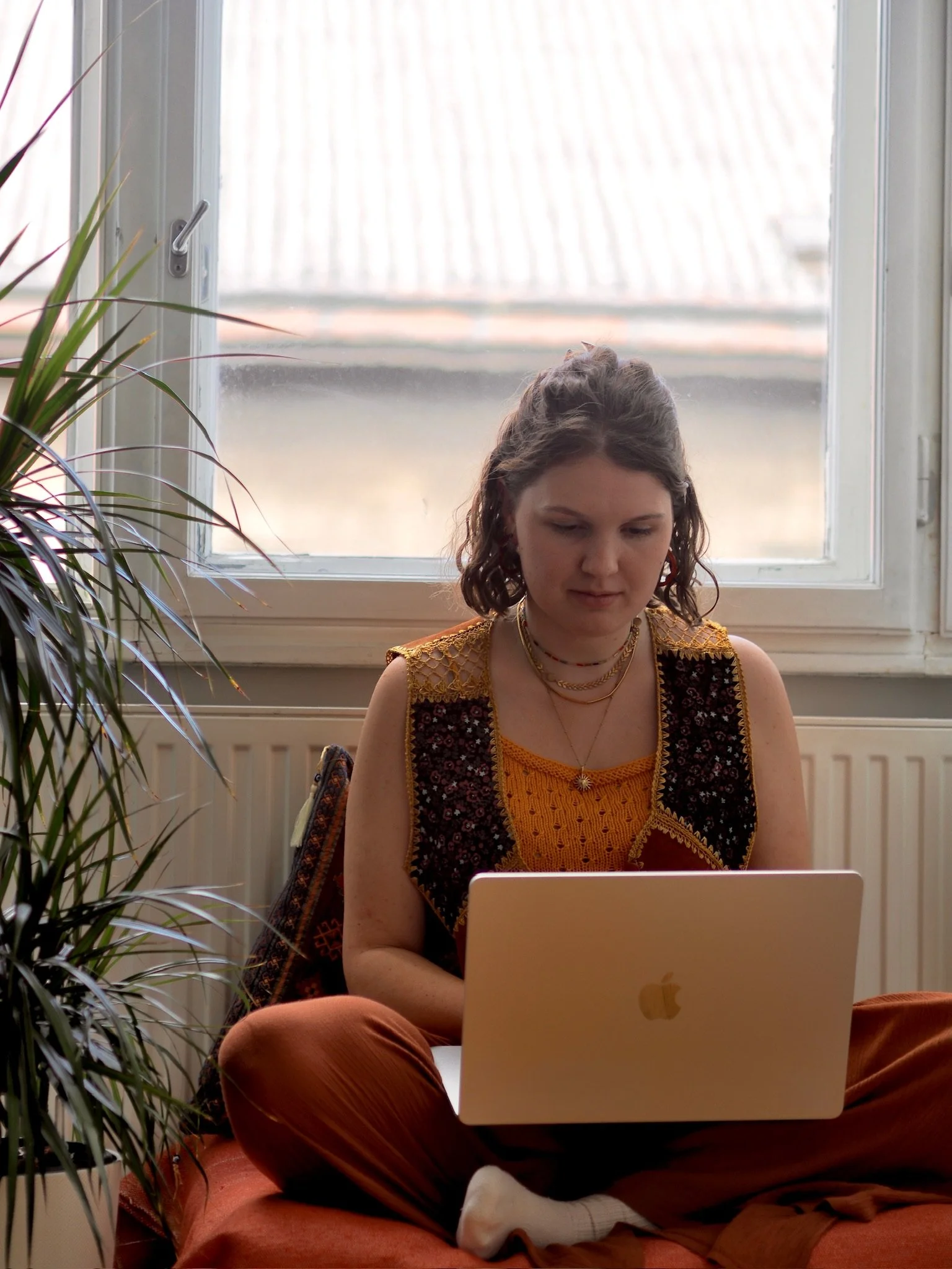 Junge Frau mit braunen, gewellten Haaren sitzt auf einem roten Sofa vor einem Fenster und arbeitet an einem Laptop, neben ihr steht eine grüne Zimmerpflanze.