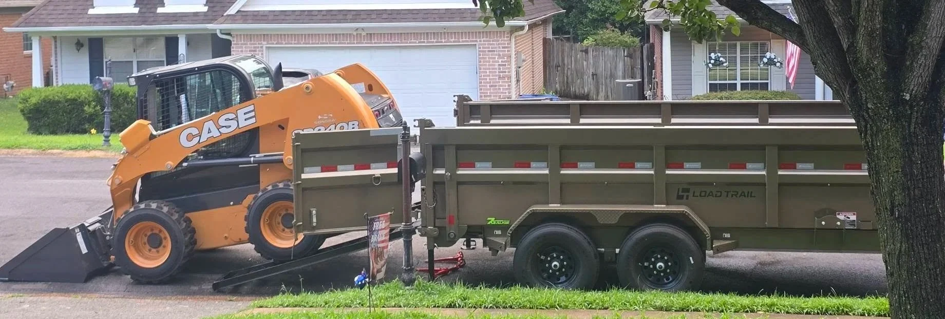 A Kubota mini skid steer loader attached to a Load Trail flatbed trailer parked on a residential street in front of a house with a brick garage and a mailbox. A tree partially covers the right side of the image.