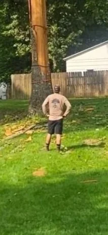 Man standing in a backyard looking at a large tree with a climbing harness attached to the trunk.
