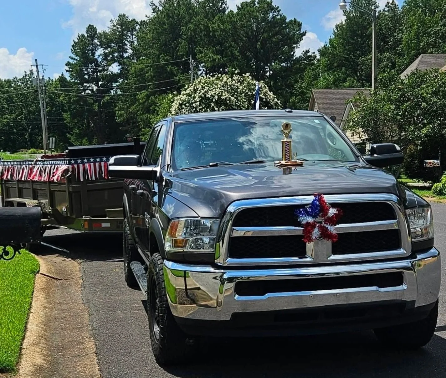 A black pickup truck decorated with patriotic red, white, and blue garland on the front grille, parked on a suburban street. A trophy is placed on the hood. The truck has a trailer attached with patriotic bunting draped along its side.