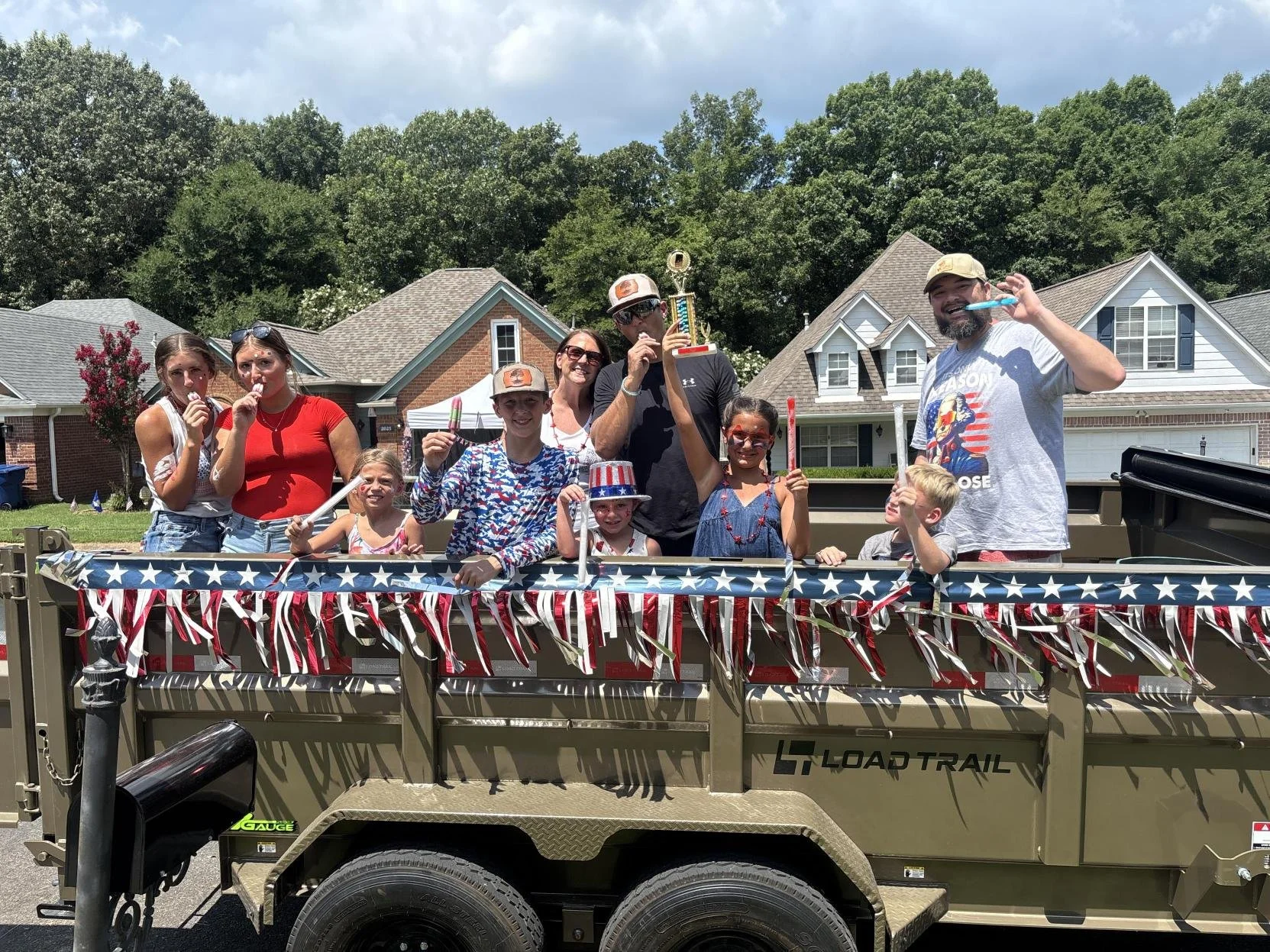A group of children and adults celebrating on a decorated float with American flags, holding sparklers, and some wearing patriotic accessories like a Uncle Sam hat and red, white, and blue clothing.
