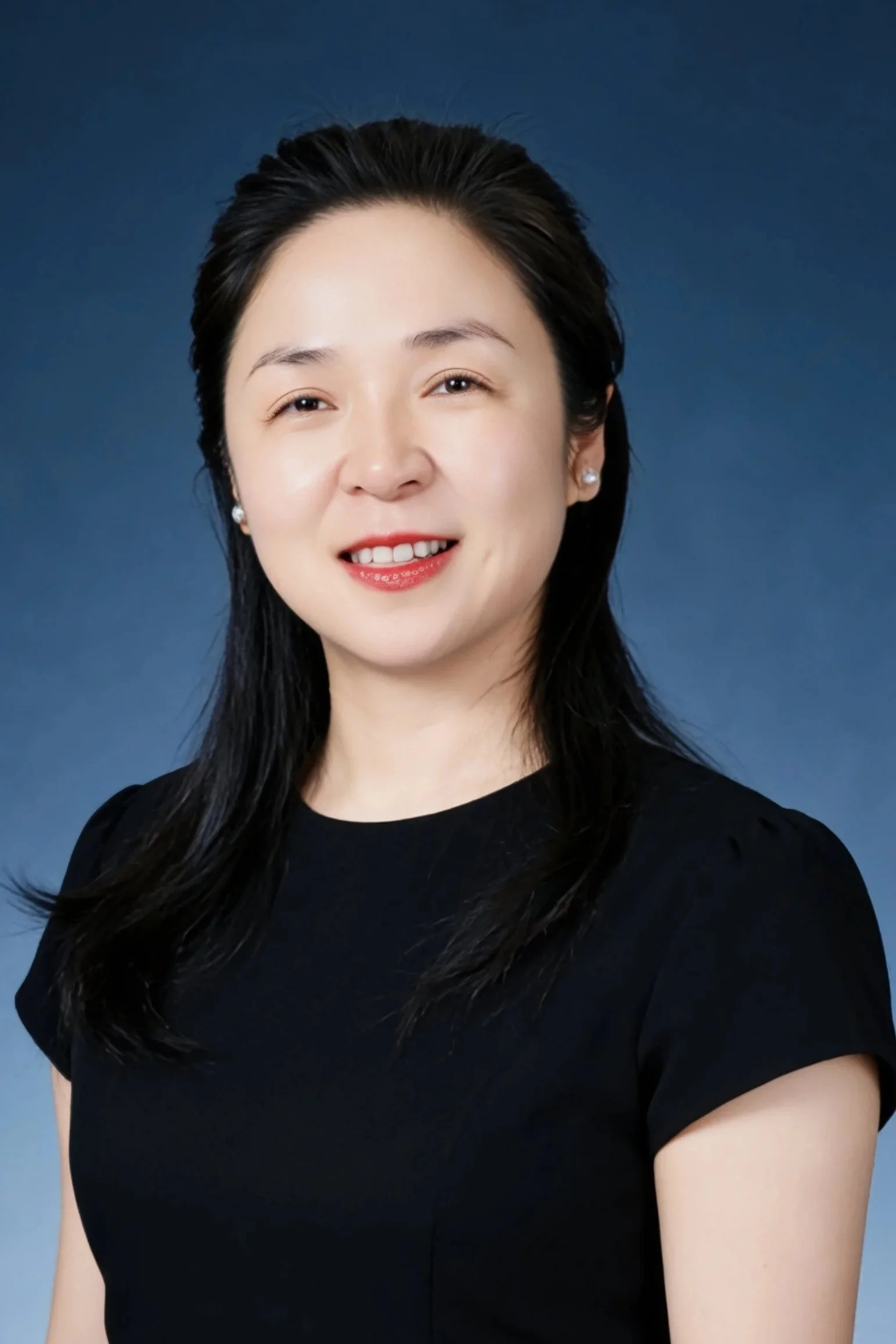 Professional portrait of a woman with black hair, wearing a black top and pearl earrings, smiling against a dark blue background.