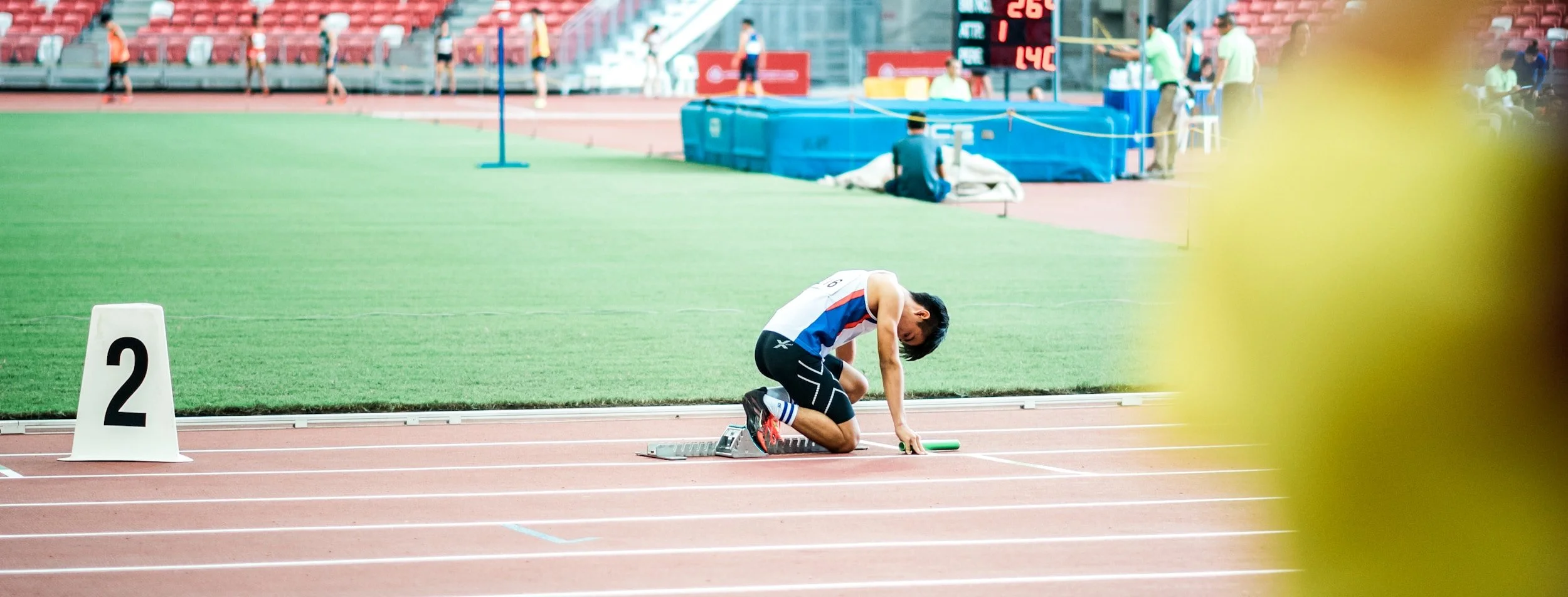 A male track athlete kneeling on a running lane at a stadium, preparing for a race with a relay baton in hand. The stadium has a green field and red track lanes with a large digital scoreboard in the background, and a few people are in the background.