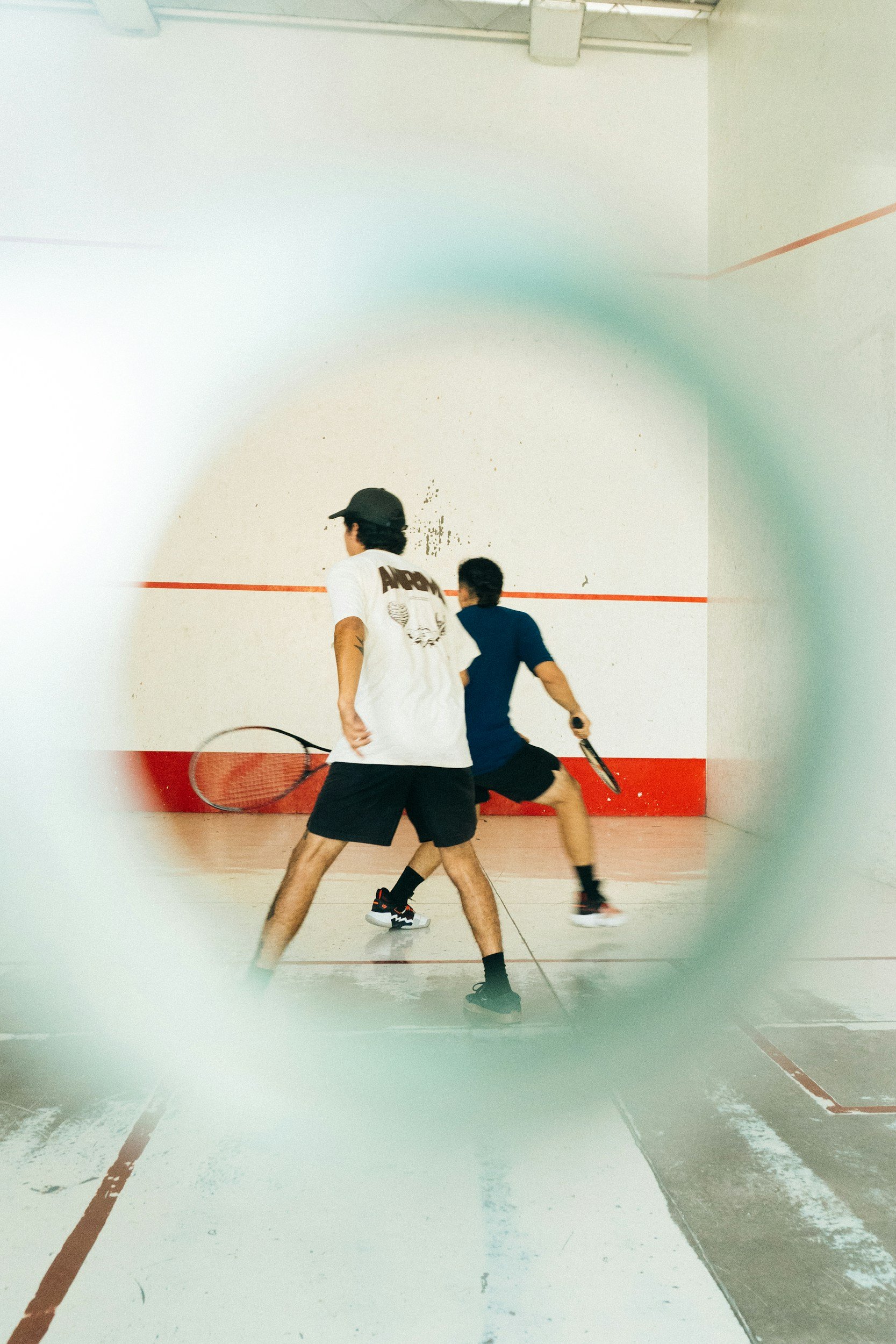 Two men playing squash on an indoor court, seen through a circular opening.