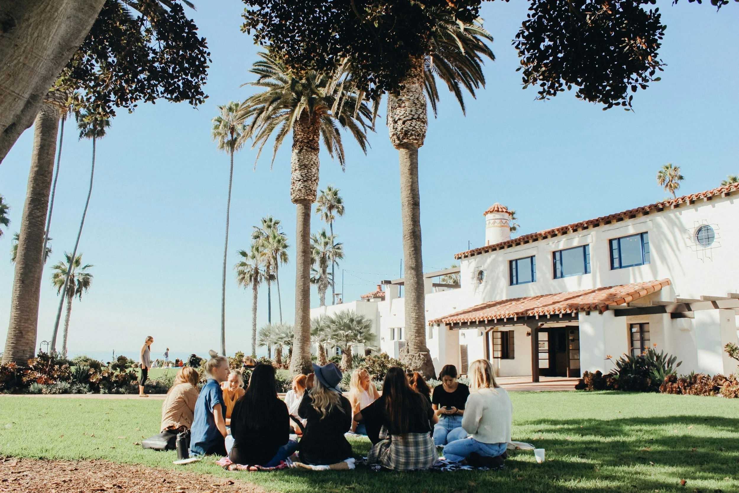 A group of people sitting on a blanket in a park with palm trees and a white Spanish-style building in the background.