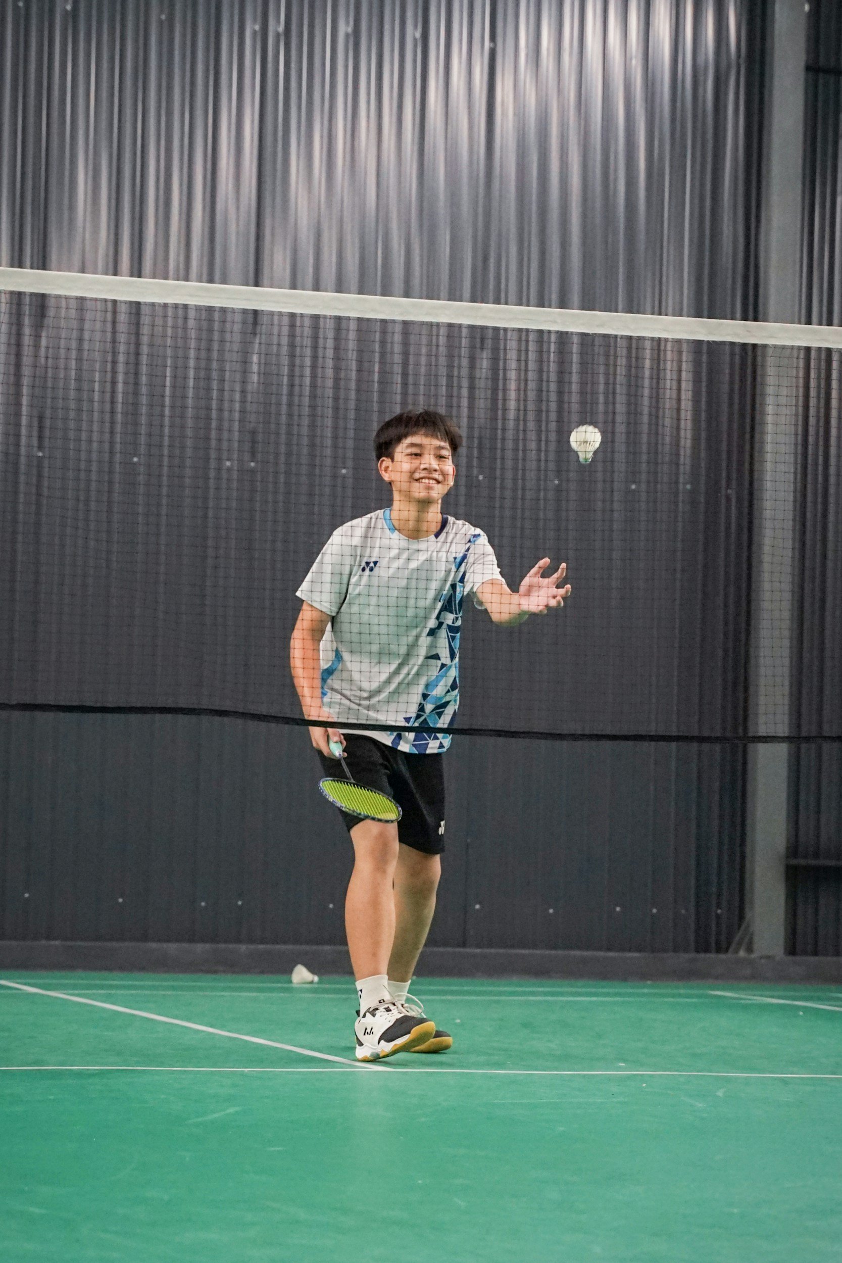 A young boy playing badminton indoors, holding a racket and reaching out to hit a shuttlecock, with a smile on his face.